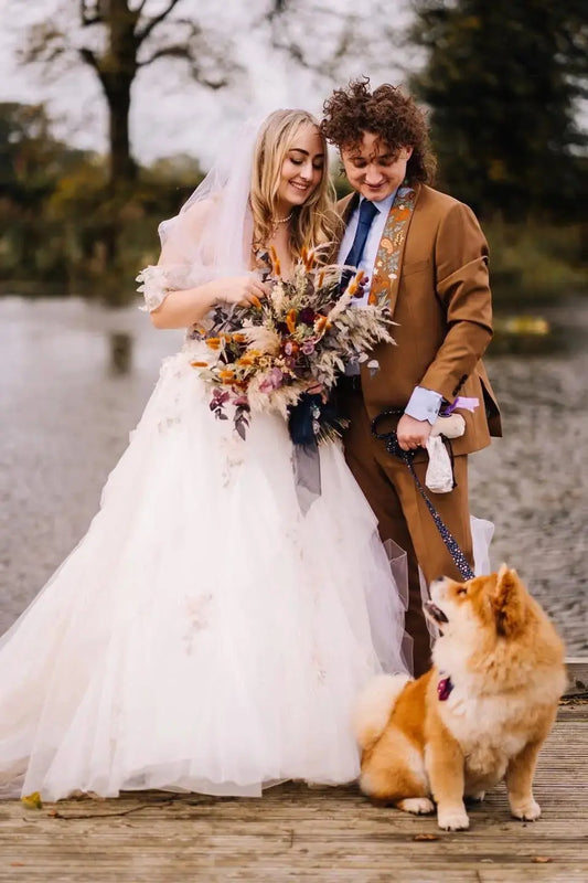 A bride, holding a large wild dried flower wedding bouquet, groom and their dog all in matching autumn colours. Stood in the grounds of a stately home  