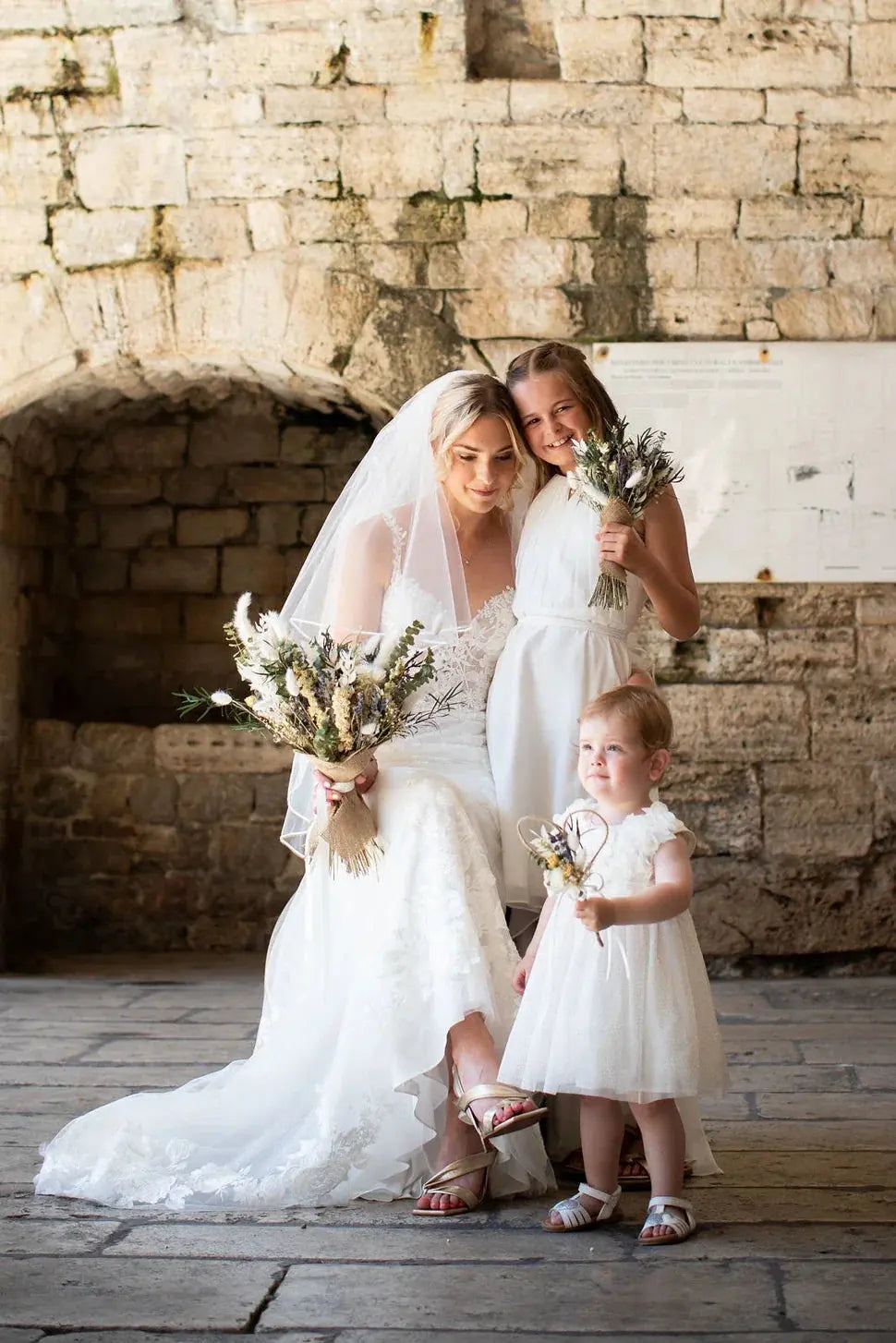 A bride, holding a large wild dried flower wedding bouquet in neutral shades. She is bent down with two flower girls in an embrace, 