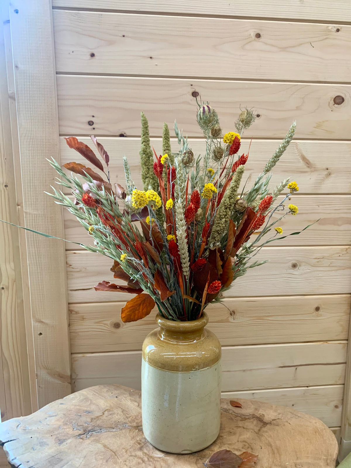A rustic dried flower arrangement in a glazed ceramic vase, set on a wooden table against a light wood plank wall.
