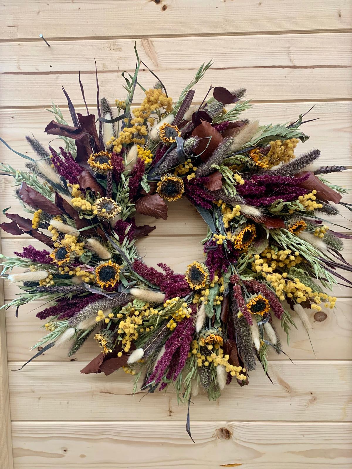 A round dried flower wreath featuring yellow mimosa, dark purple milkweed, sunflowers, and green foliage, displayed against a light wood plank wall.