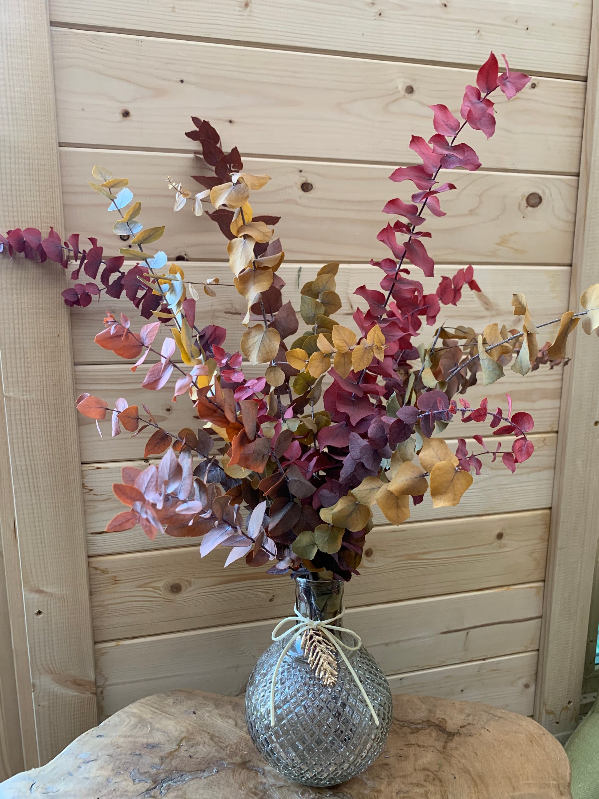A decorative arrangement of dried autumn leaves in shades of burgundy, brown, and tan, displayed in a textured gray woven vase tied with white string, set against a light wood plank background.