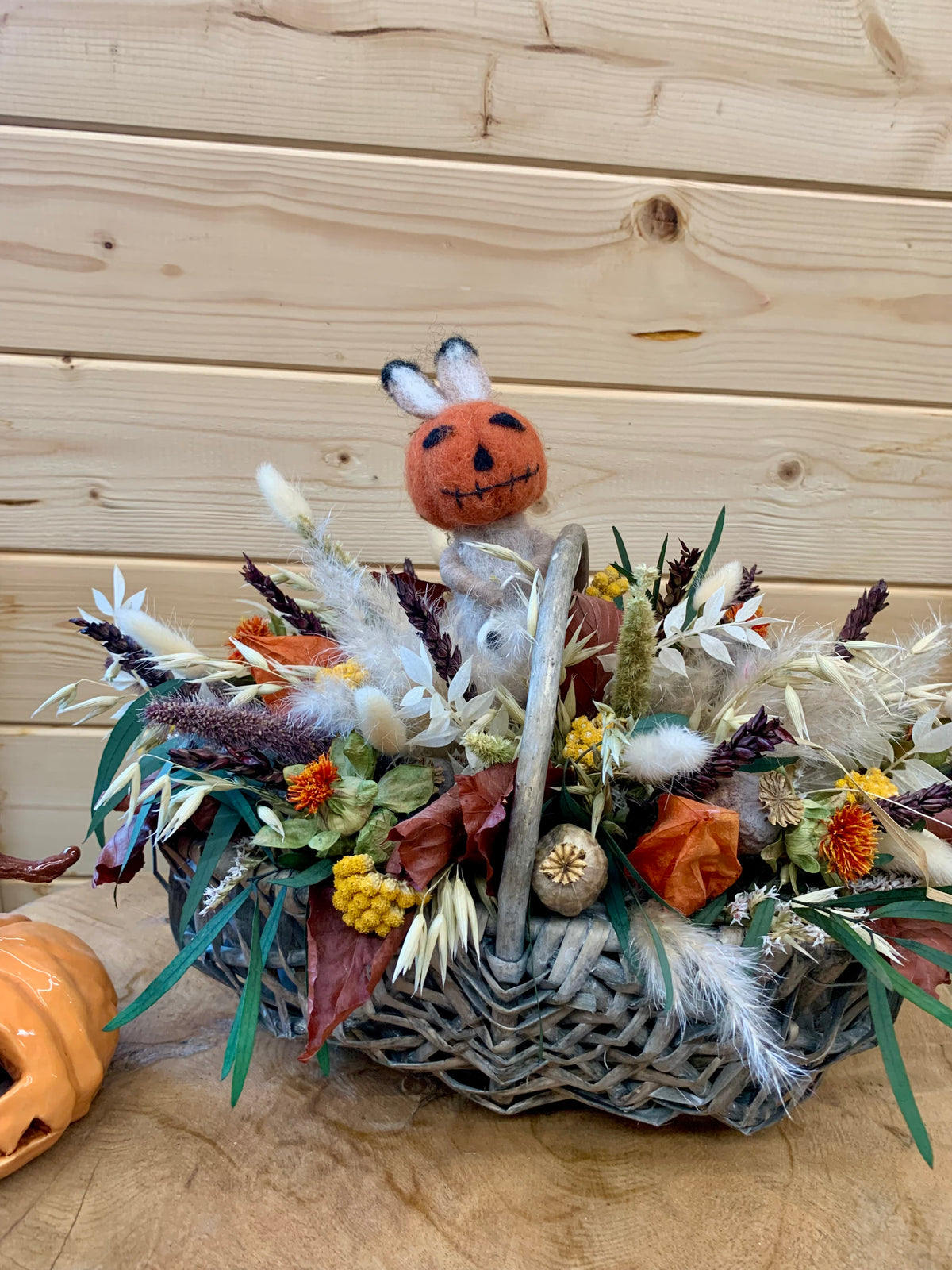 An autumn-themed woven basket centerpiece filled with dried grasses, white feathers, orange leaves, small yellow flower clusters, and a small felt jack-o'-lantern character with bunny ears, set against a light wood plank background.