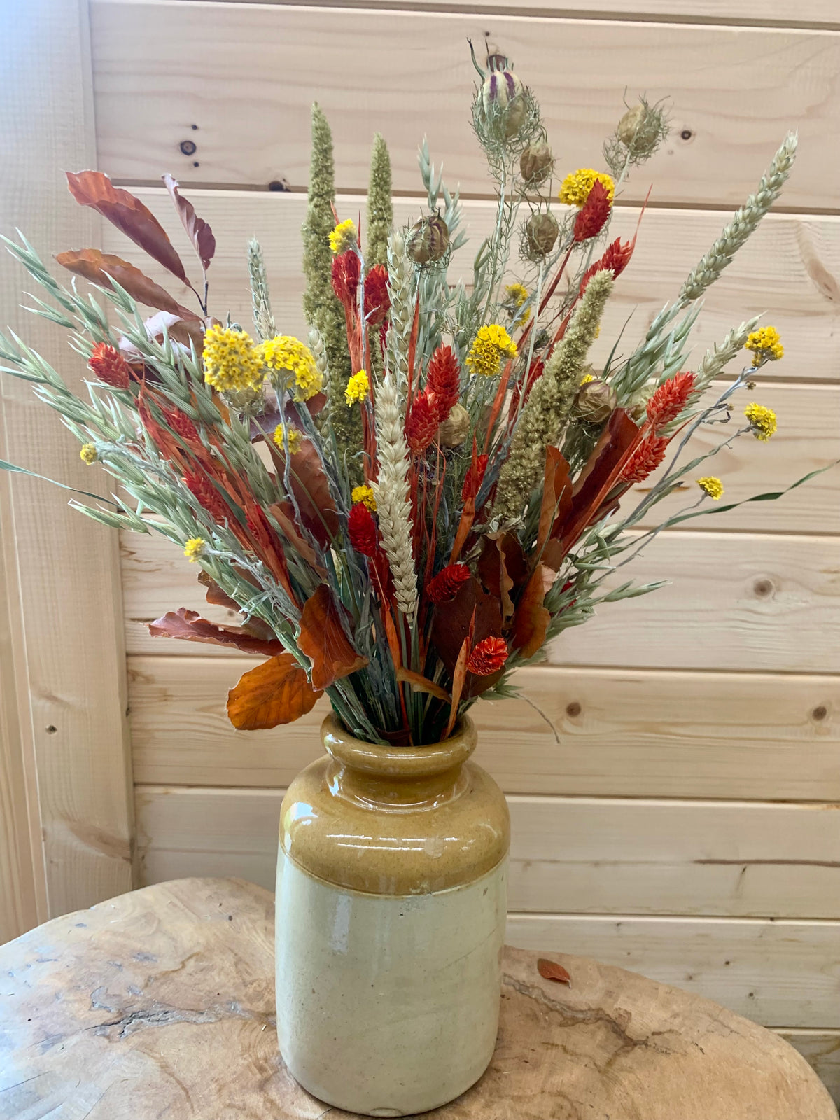 An autumnal dried flower arrangement in a rustic stoneware vase, set on a wooden table against a light wood paneled wall.