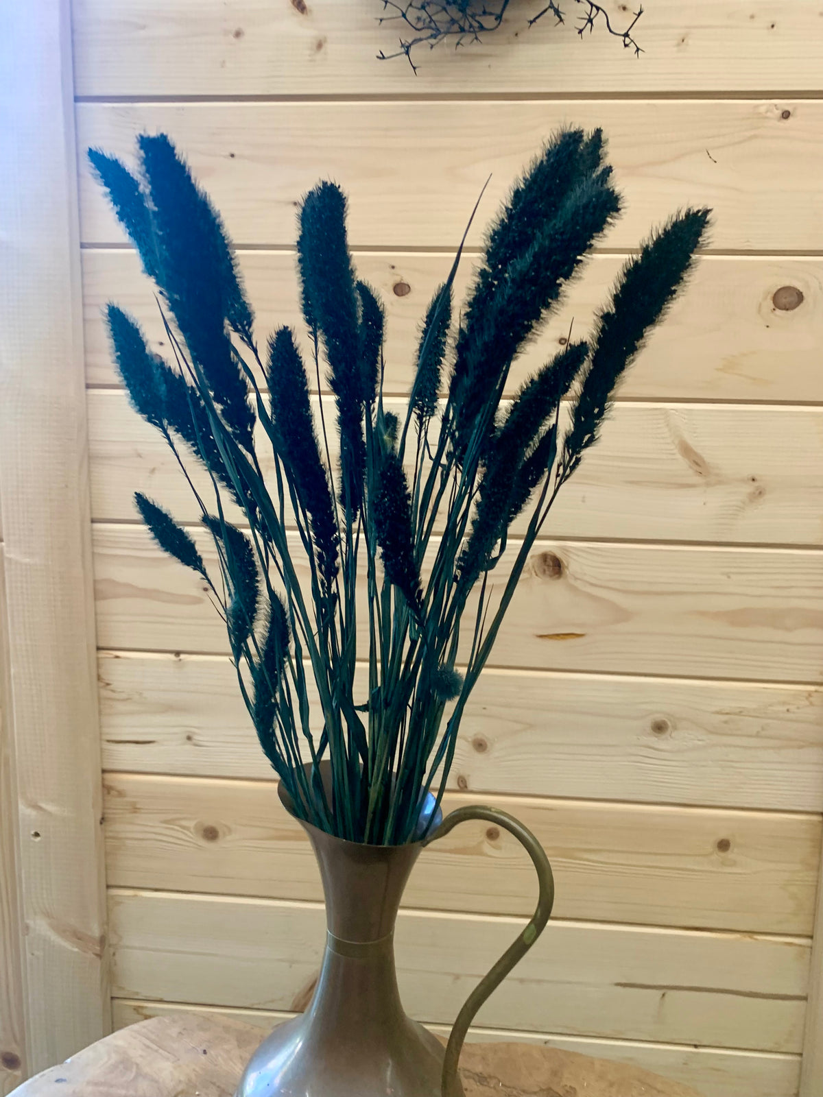 Dark green/black setaria grass stems arranged in a copper/brass pitcher vase, set against a light wood plank wall.