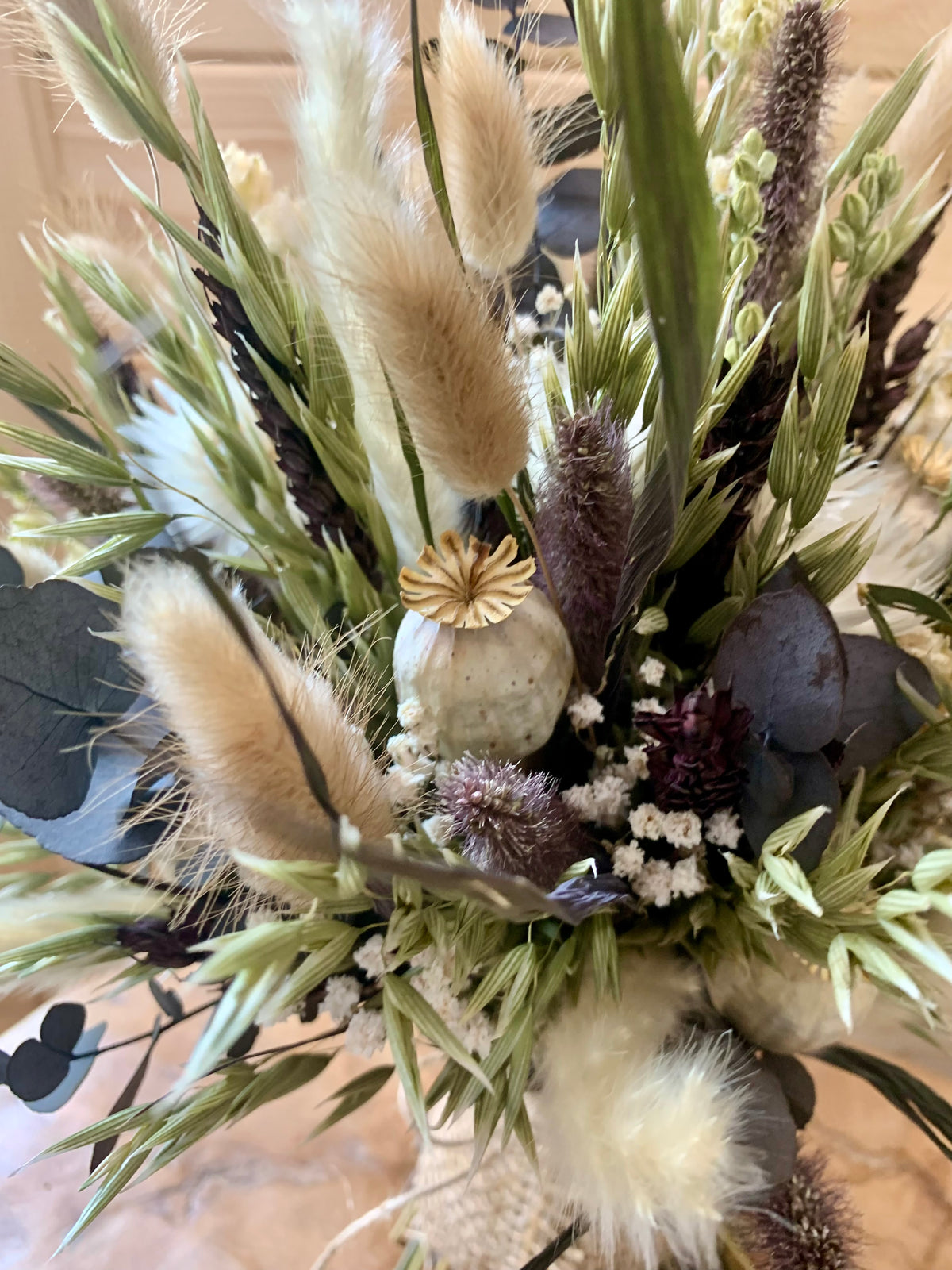 A close-up of a dried floral arrangement featuring beige bunny tail grass, green foliage, dark purple thistle-like stems, eucalyptus leaves, and a small pale stone vase with a gold flower accent.