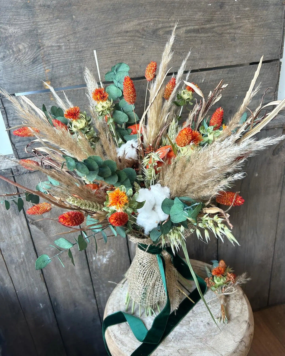 Dried flower bouquet using forest greens, neutral pampas grass and orange flowers finished with forest green velvet ribbon on a wooden surface with a matching buttonhole.