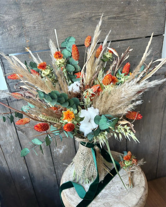 Dried flower bouquet using forest greens, neutral pampas grass and orange flowers finished with forest green velvet ribbon on a wooden surface with a matching buttonhole.