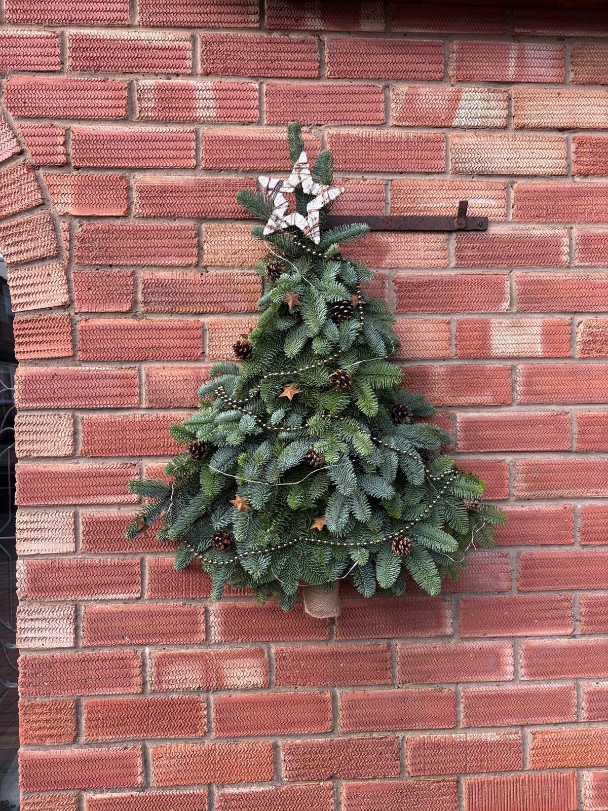 A decorative fresh Christmas tree wall hanging made of layered green pine branches, with small pinecones, star ornaments, and a white star topper, mounted against a red brick wall.