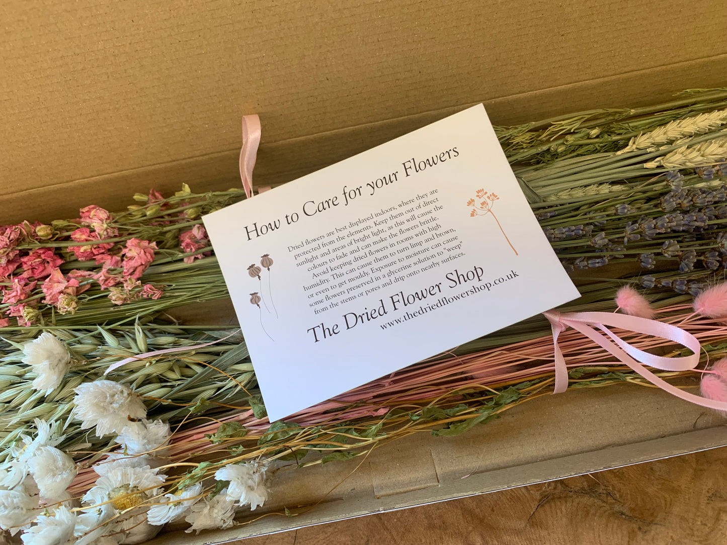 A gift box of dried flowers and greenery with a white care card from 'The Dried Flower Shop' resting on top. The arrangement includes pink, white, and purple dried blossoms with pink ribbons tied around it.