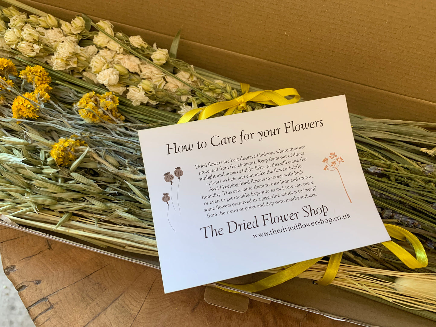 A white printed care card titled 'How to Care for your Flowers' from 'The Dried Flower Shop' placed on top of a bundle of dried flowers inside a cardboard box. The flowers include cream, yellow, and lavender tones, tied with a yellow ribbon.