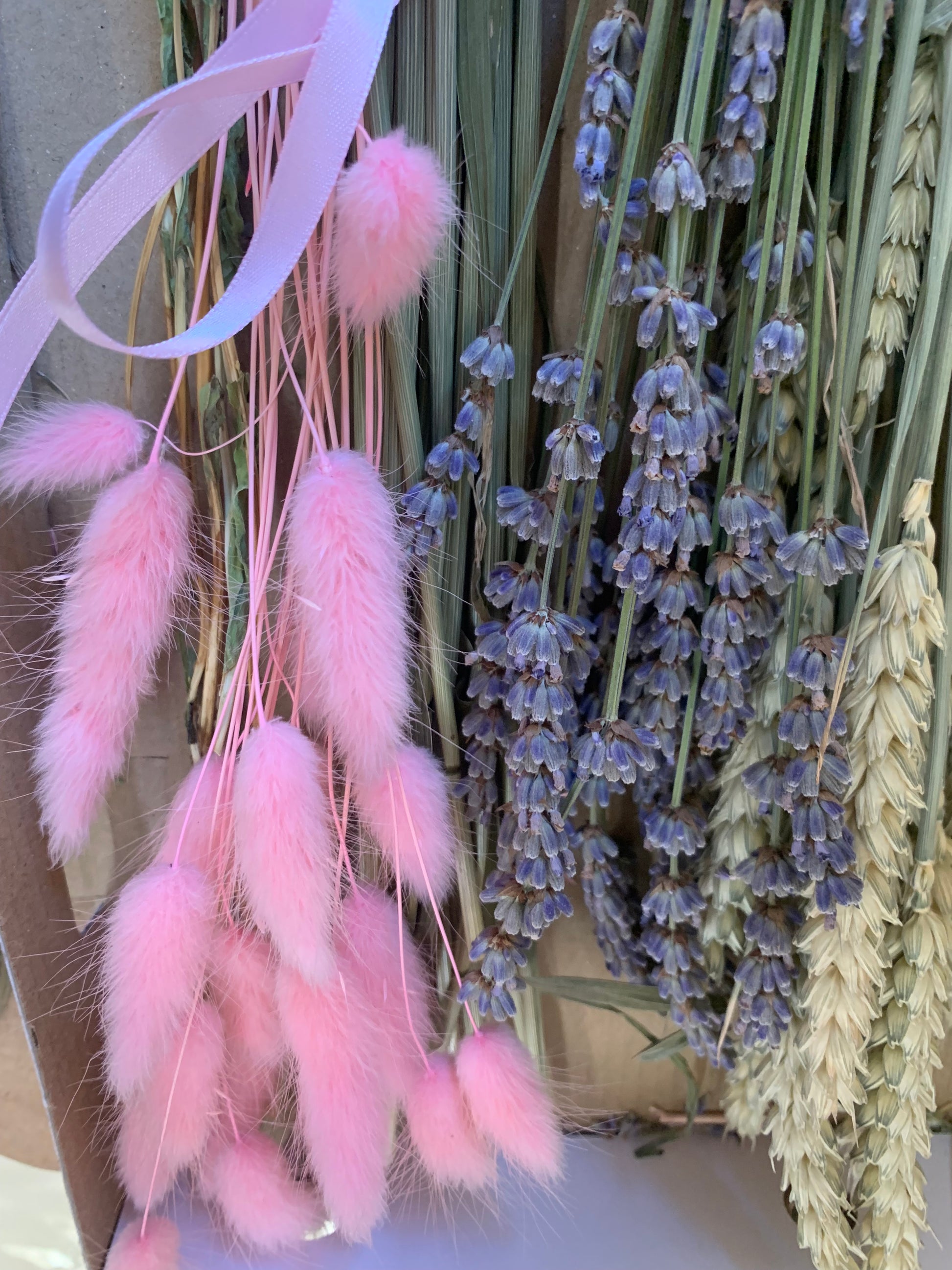 A bundle of decorative dried flowers and wheat stems, including pink bunny tail grass, lavender, and golden wheat, arranged together with a pale pink ribbon.