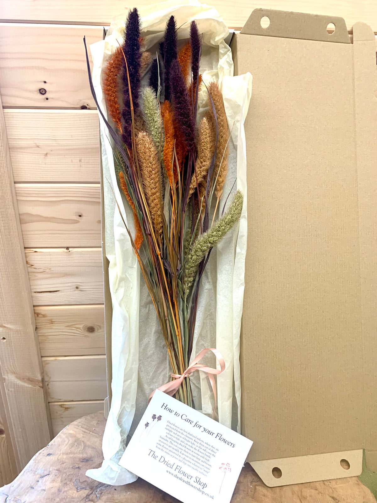 A bundle of dried decorative grasses wrapped in white tissue paper and tied with a pale peach ribbon, with a printed care card in front. The bouquet is photographed indoors against a light wood paneled wall and in a cardboard box.