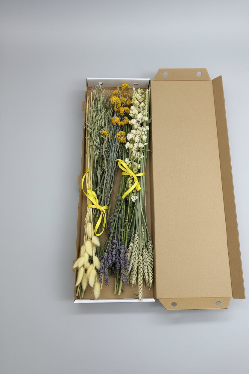 A dried flower and botanical gift box containing white baby's breath, yellow flowers, wheat, lavender, and green stems tied with yellow ribbons, shown inside an open brown cardboard box.