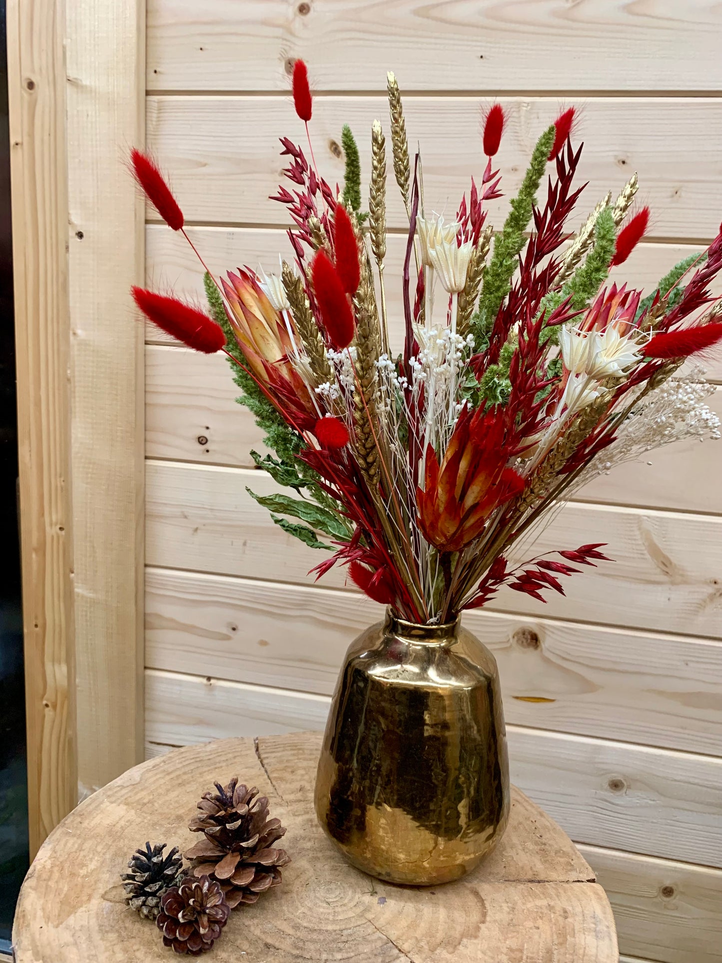 Gold vase with festive dried flowers and pinecones on a wooden surface against a wooden wall.