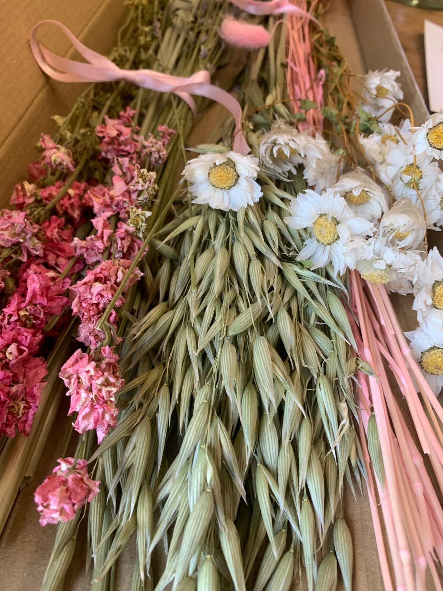 A close-up of dried flower bundles arranged in a box, featuring pink blossoms, white daisy-like flowers with yellow centers, and pale green grass-like stems tied with a light pink ribbon.