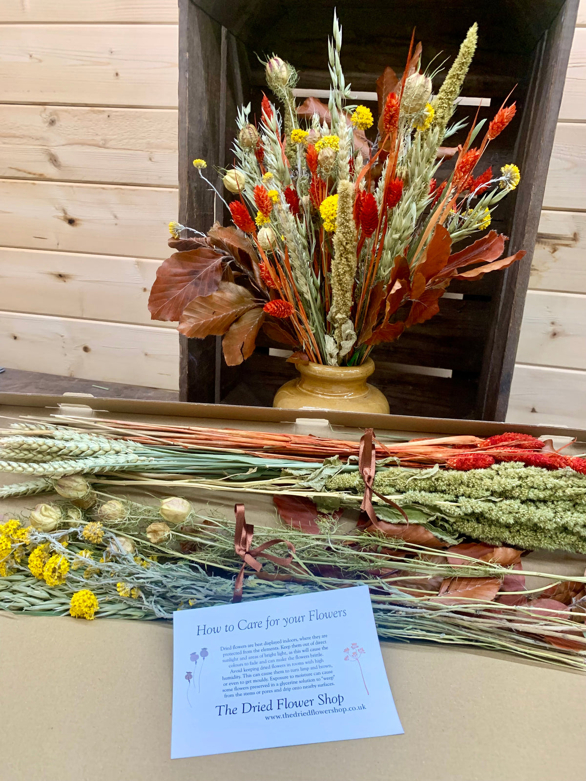 A dried flower arrangement in a small beige ceramic pot displayed on a table, with additional loose dried flowers and a care card in front. The arrangement features wheat, red berries, yellow flowers, and brown leaves.