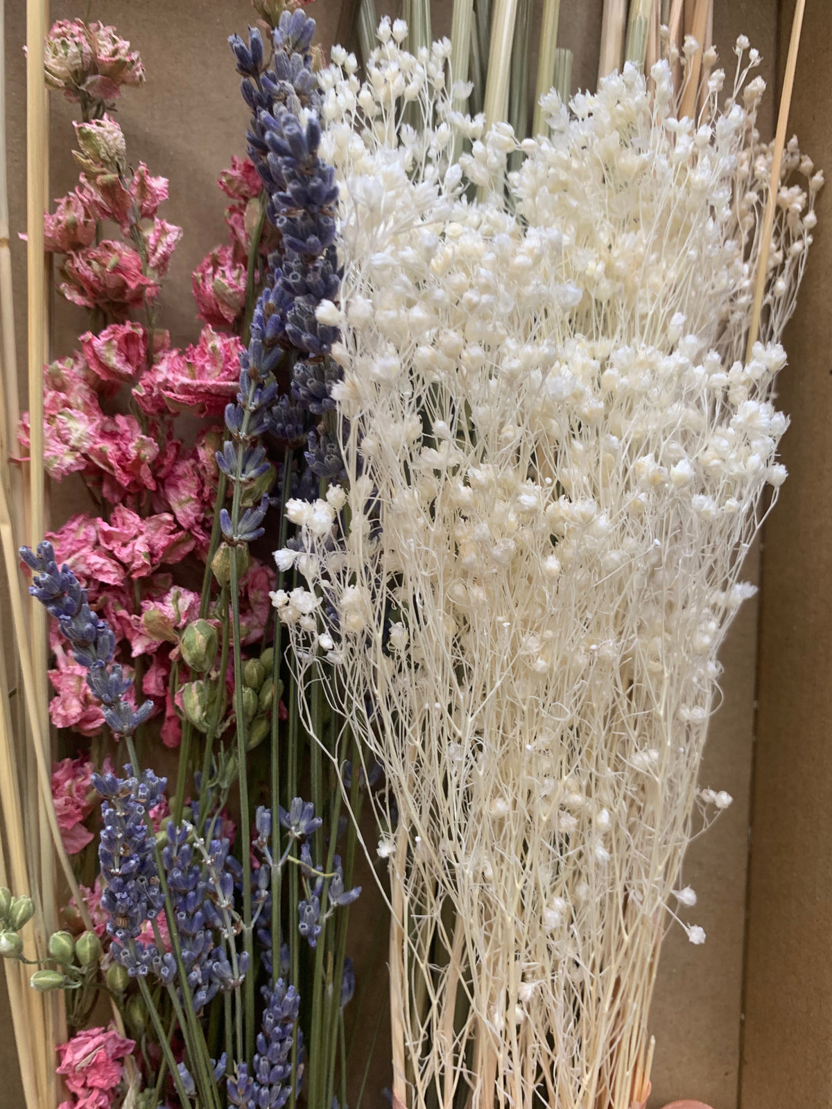 A bundle of dried flowers including white baby's breath, purple lavender, and pink larkspur blossoms arranged against a brown cardboard background.