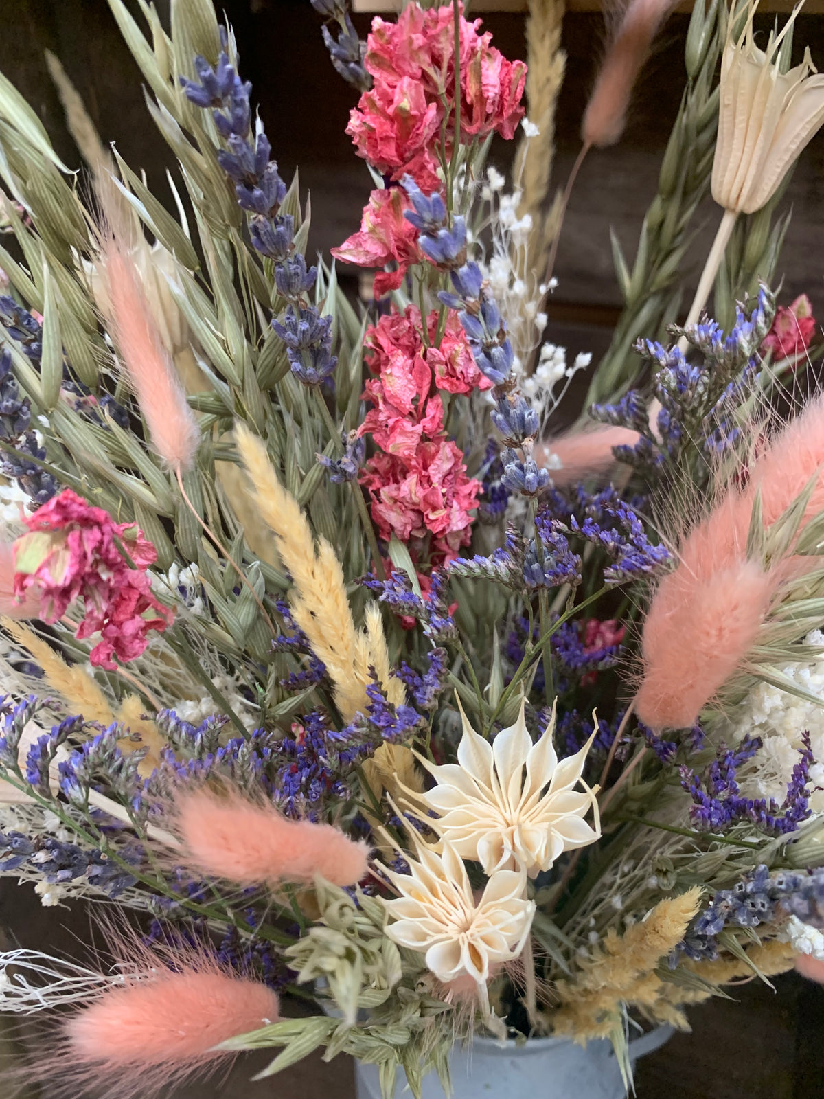 A dried flower arrangement featuring pink blossoms, lavender sprigs, beige wheat-like stems, and fluffy blush bunny tail grass, arranged in a metal container.