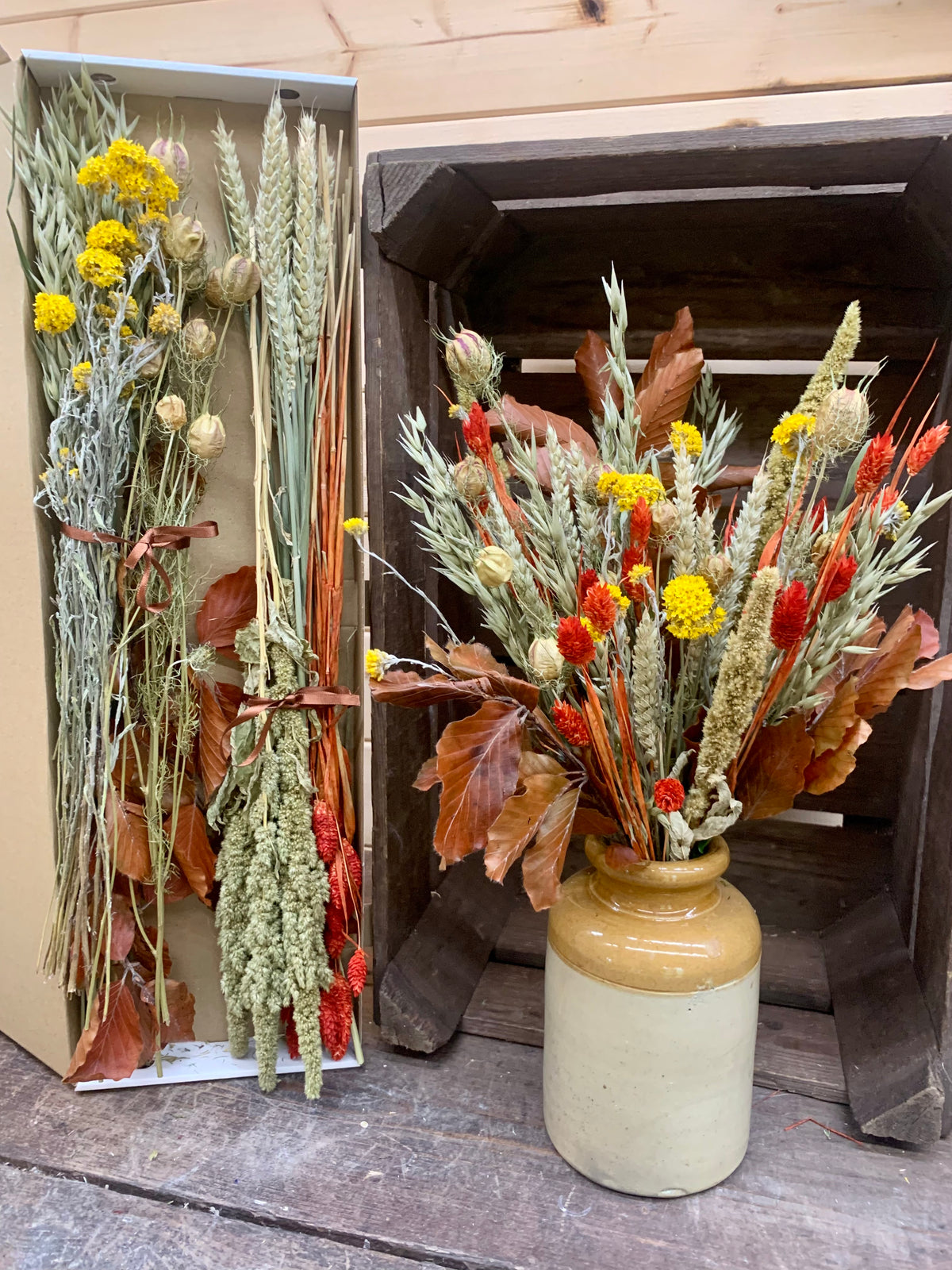 A dried flower arrangement displayed in a rustic setting, featuring a ceramic vase with a yellow and beige glaze on the right, and a matching rectangular box with dried botanicals on the left, set against a dark wooden crate and light wood background.