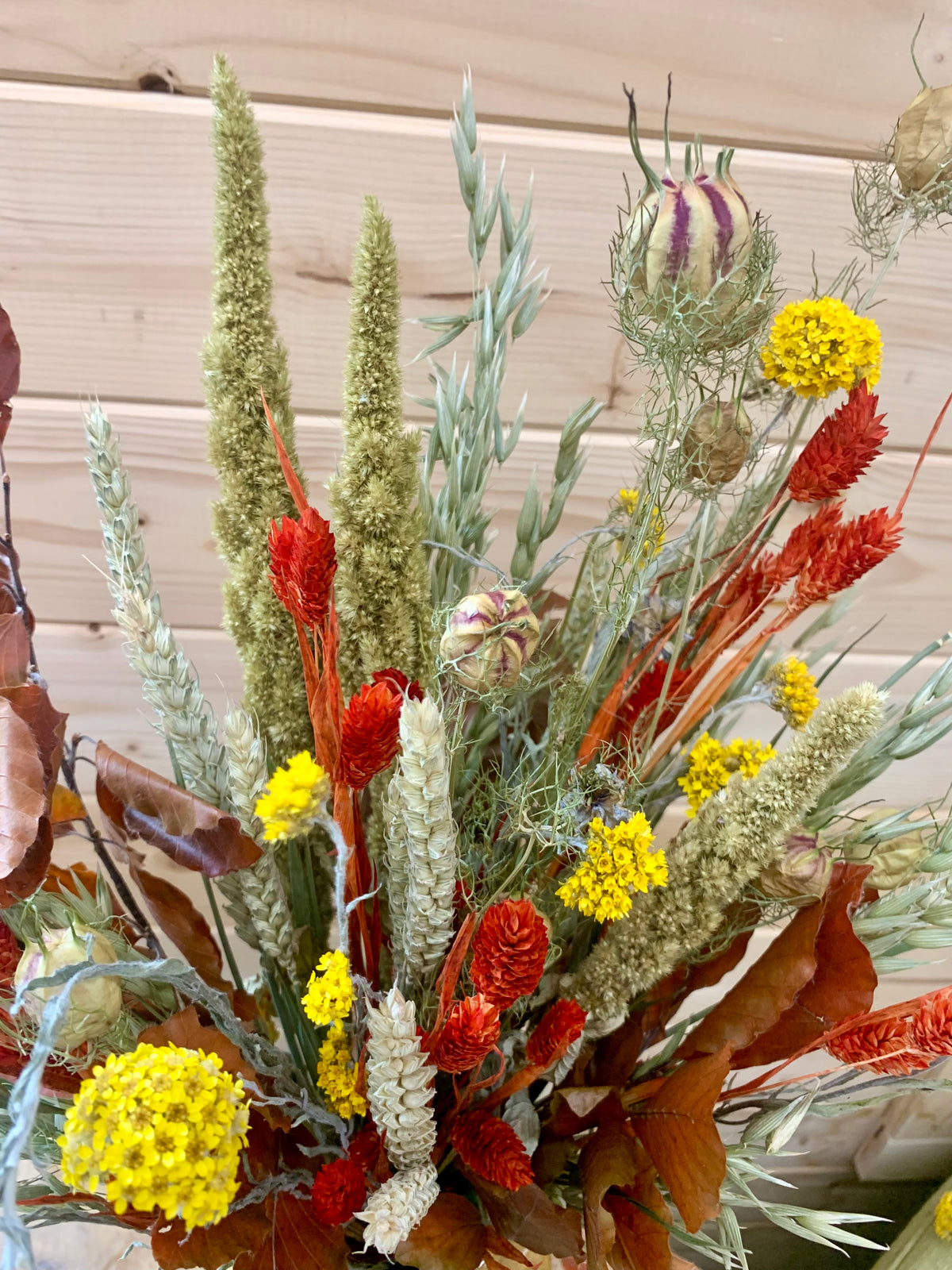 A dried floral arrangement featuring tall beige and green grasses, bright yellow flowers, red-orange spikes, and brown leaves, set against a light wood plank background.