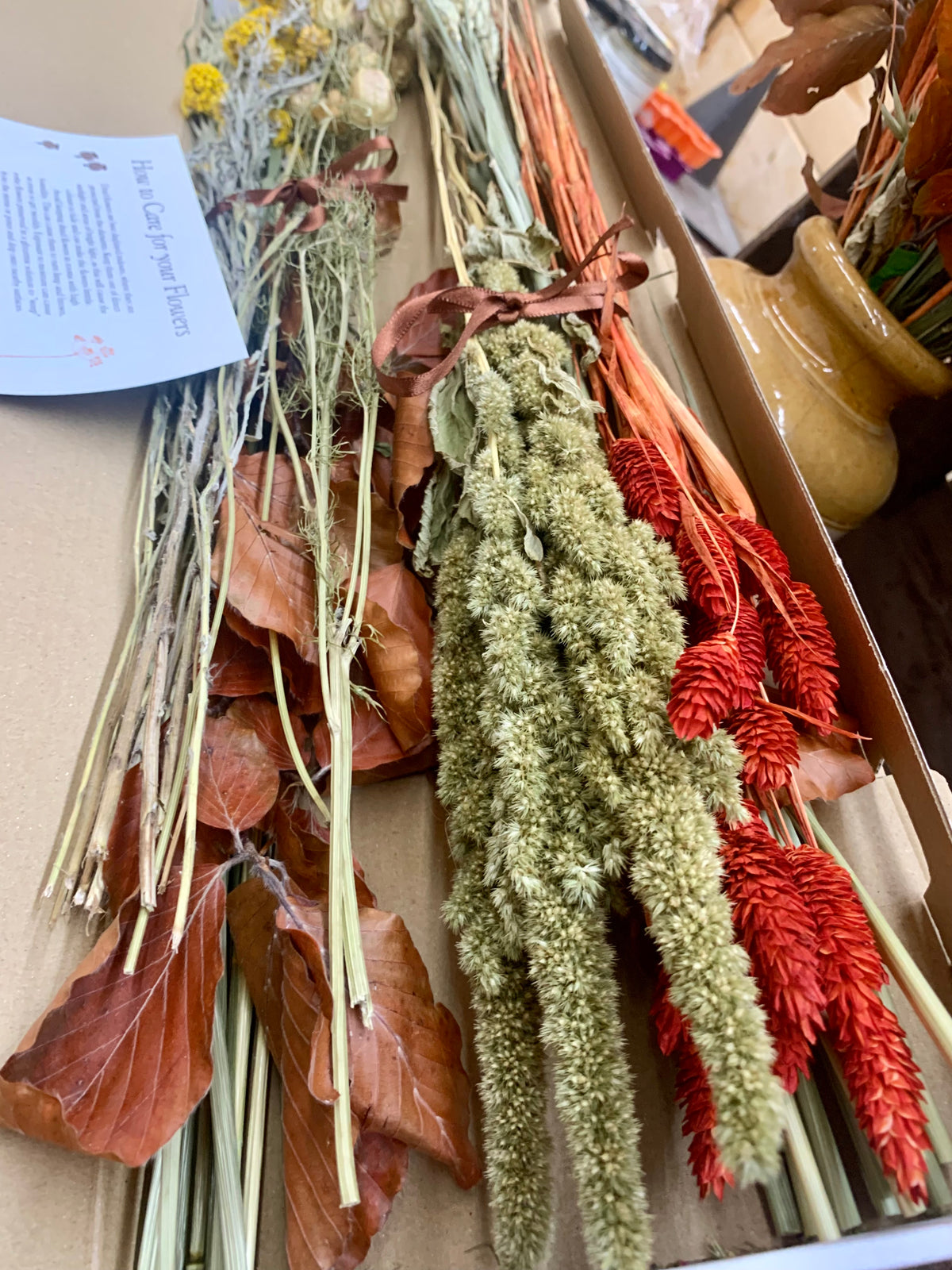 A box of dried botanicals and flower stems, including tan and rust leaves, pale green seed heads, and bright red ornamental grasses, photographed inside an open cardboard package.