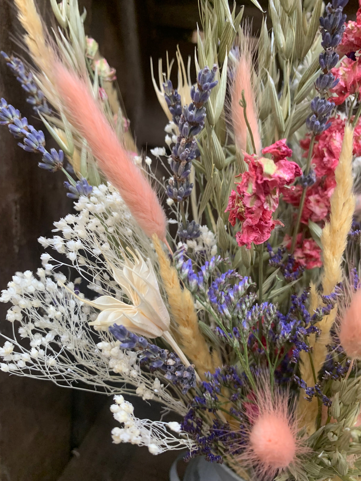 A close-up of a dried flower arrangement featuring lavender, pink ornamental grass, white baby's breath, and muted green foliage.