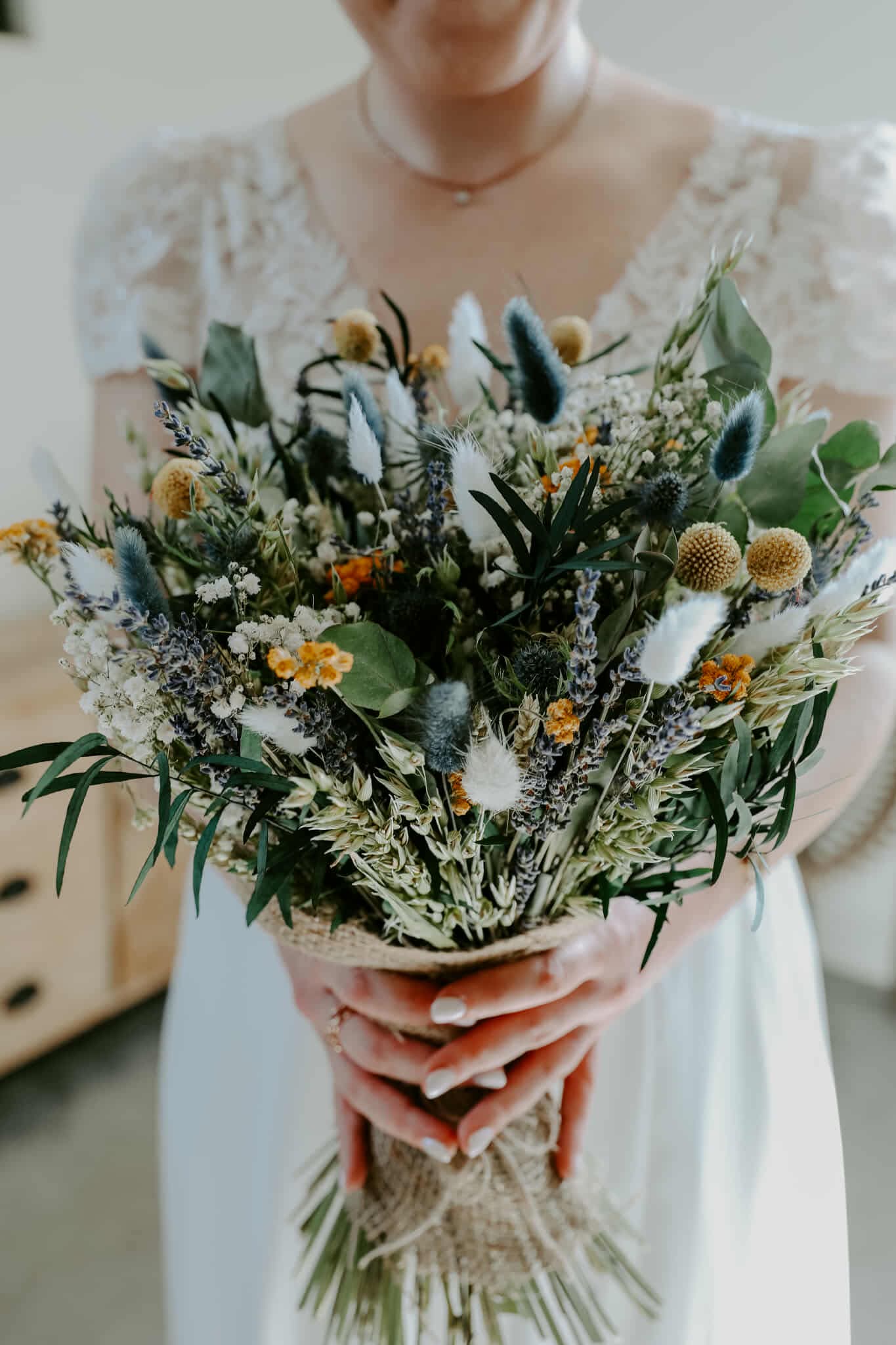 A bride holding a bouquet, of forest green, sage, blue and white dried flowers with a pop of yellow, in a softly blurred indoor setting
