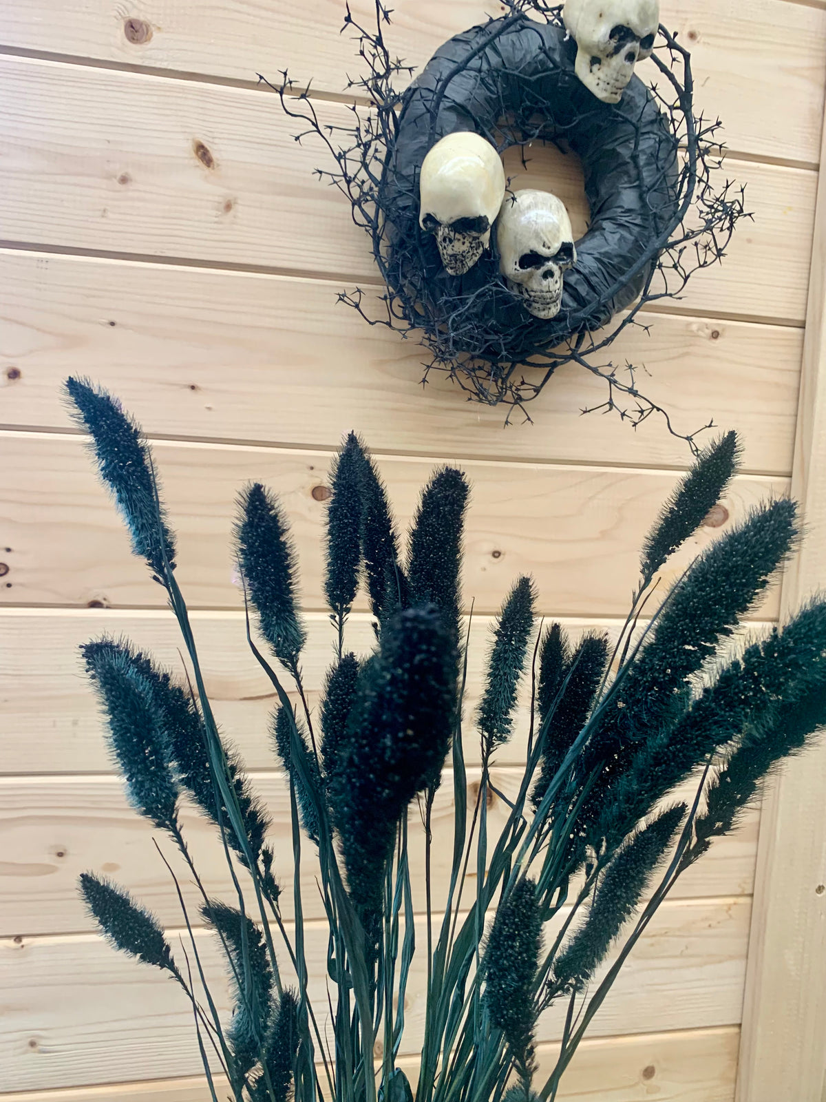 A decorative arrangement of dark green-black foxtail grass stems in the foreground, with a black wreath hanging on a light wood plank wall above, adorned with three cream-colored skulls and twisted black branches.