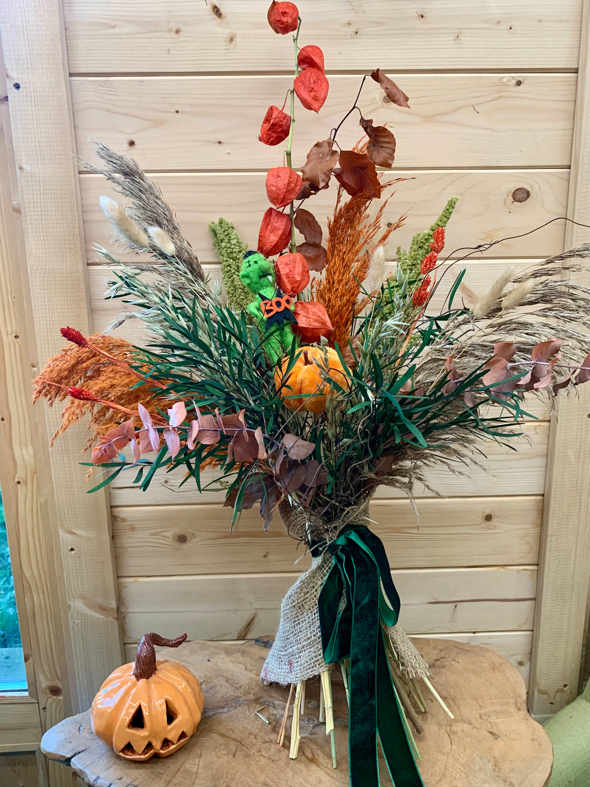 An autumn-themed floral arrangement featuring orange pumpkins, dried pampas grass, green foliage, and red leaves, tied with a dark green ribbon and displayed on a wooden stool in front of a light wood paneled wall.