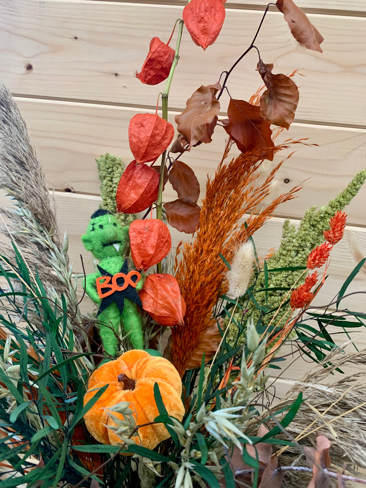 An autumn-themed decorative arrangement featuring a small green plush monster holding an orange 'BOO' sign, alongside a faux orange pumpkin, dried red berries, brown leaves, and pampas grass against a light wood plank background.