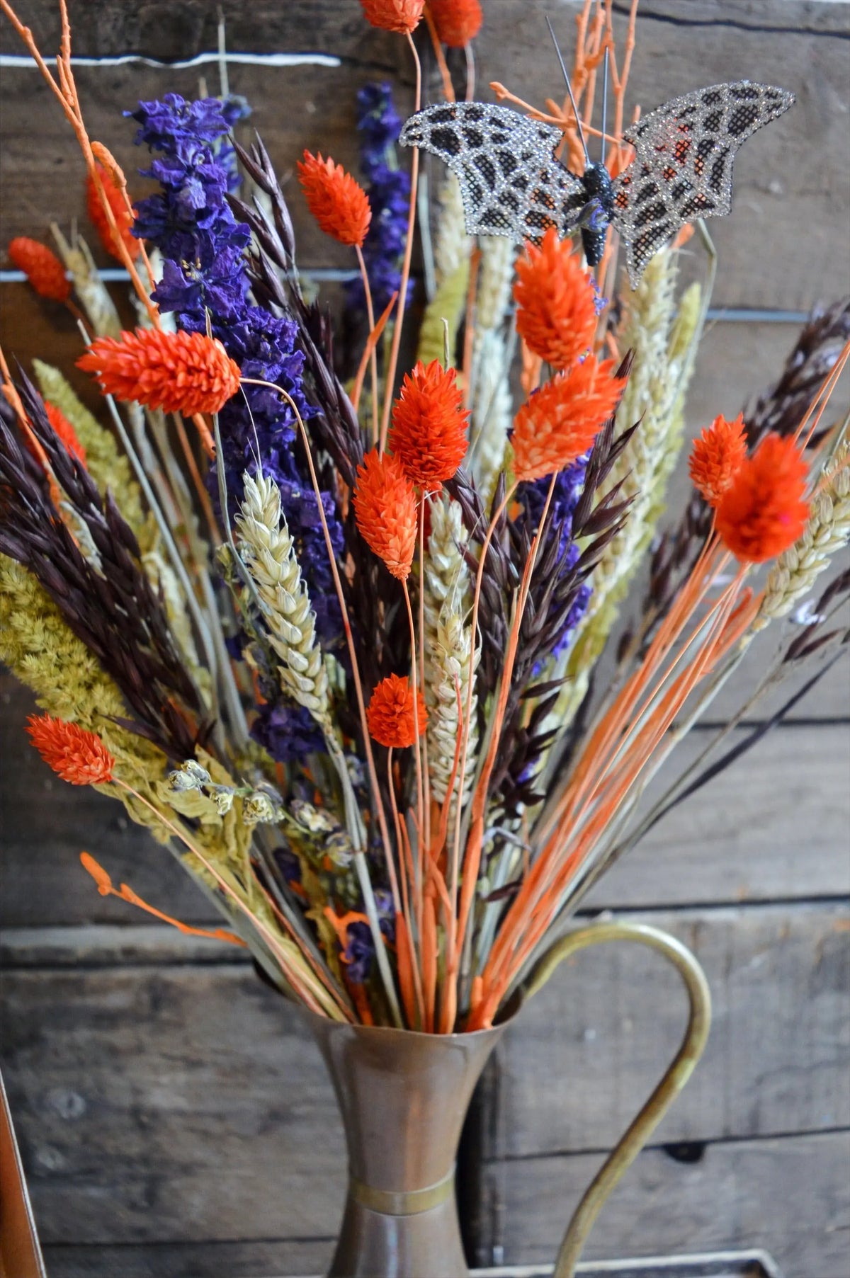 Bouquet of dried flowers in a halloween theme with orange and purple elements in a metal vase against a wooden background.