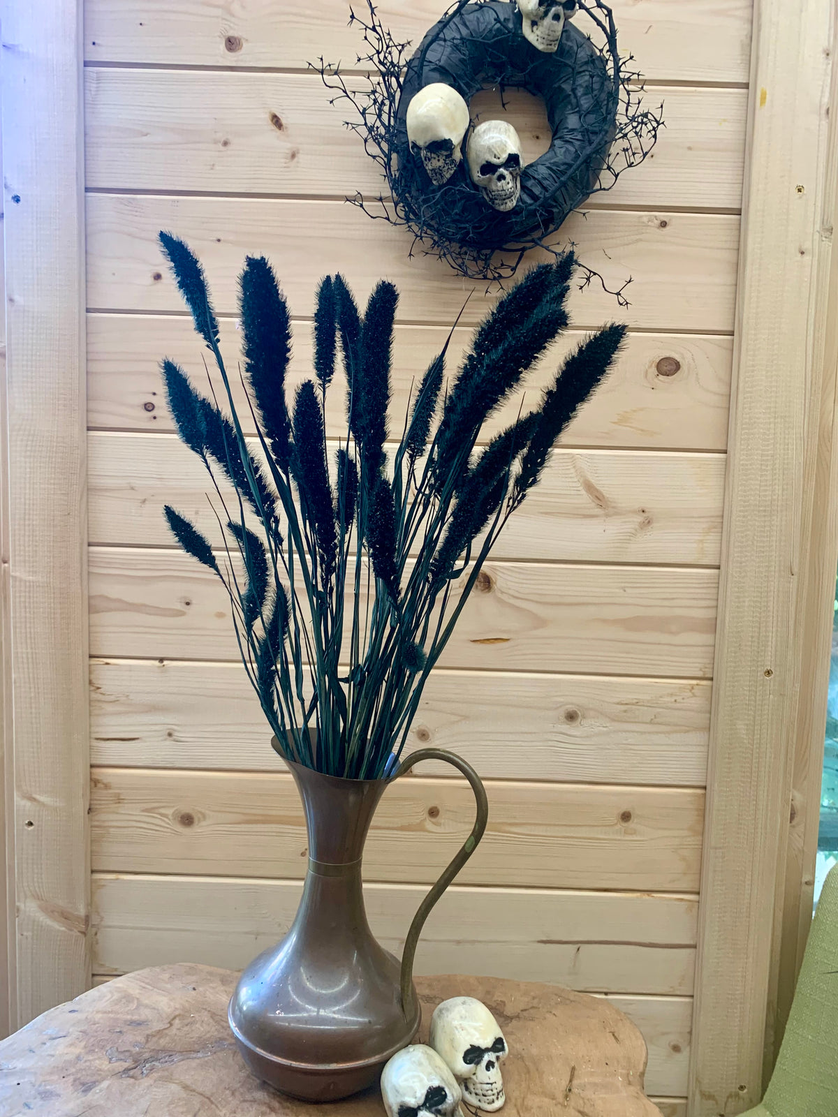 A decorative arrangement of tall black ornamental grass stems in a small metallic vase, displayed on a stone surface against a light wood plank wall, with skull decor and a wreath above.
