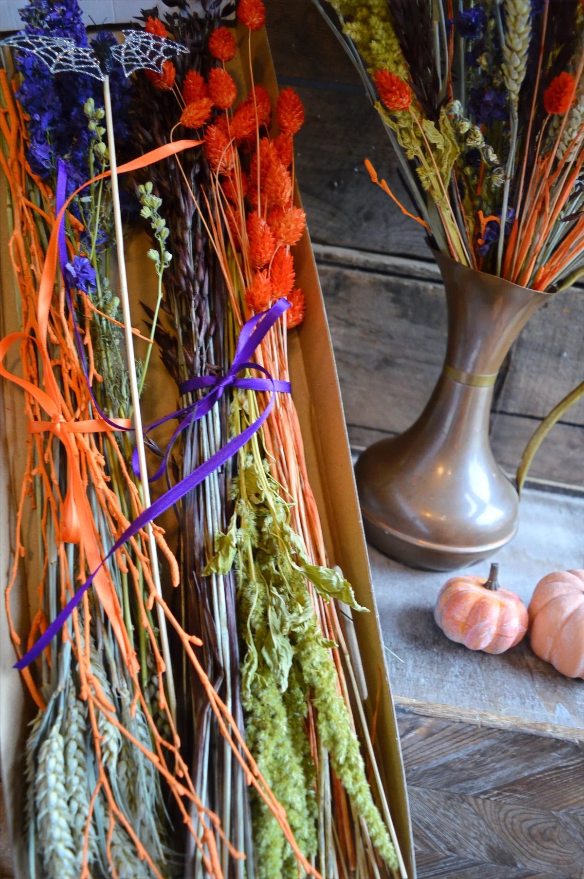 Dried floral arrangements with orange and purple ribbons in a vase on a wooden surface.