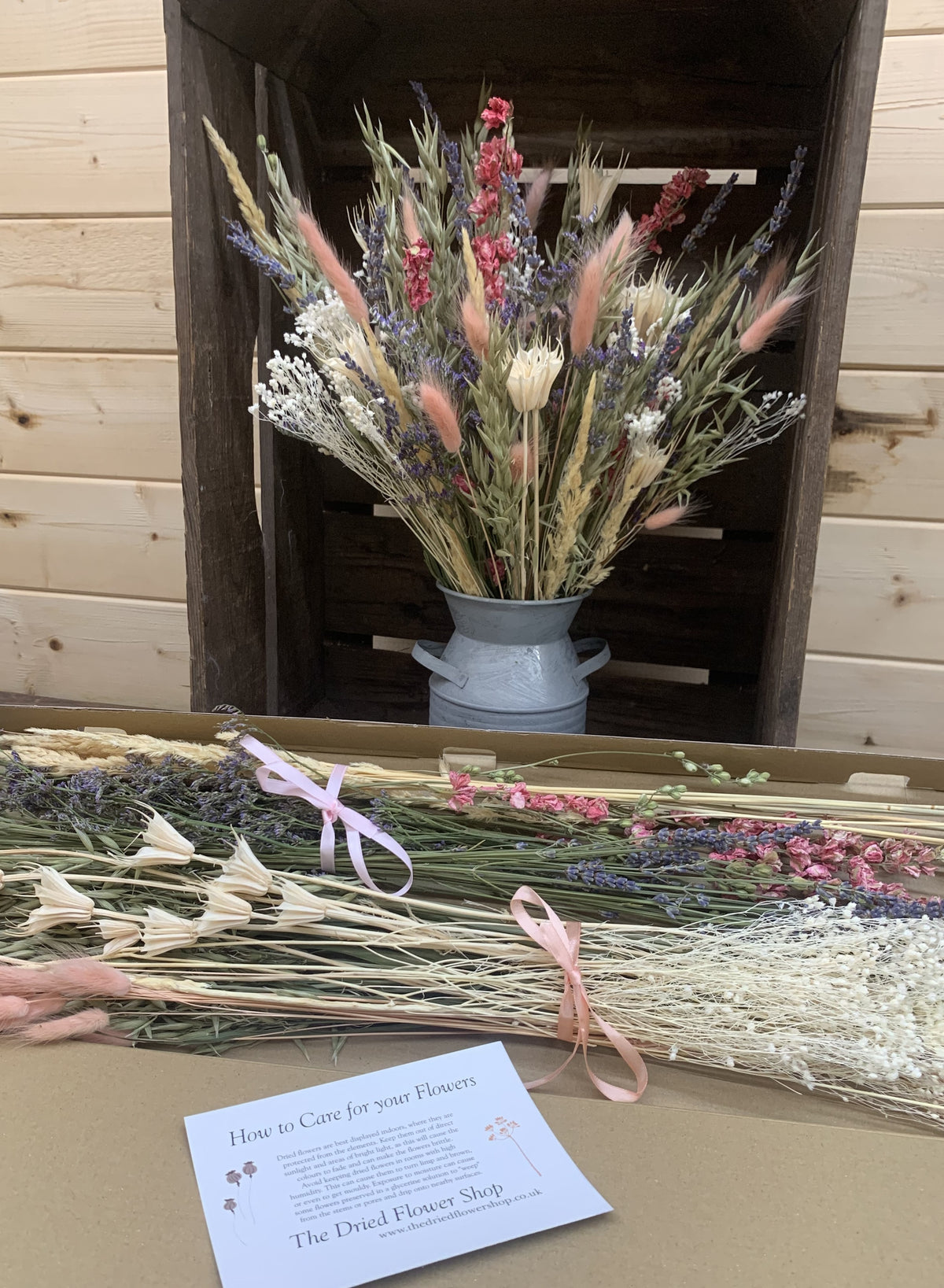 A dried flower arrangement in a small grey metal churn vase displayed inside a rustic wooden crate, with additional loose dried flowers and a care card in the foreground on a tabletop.