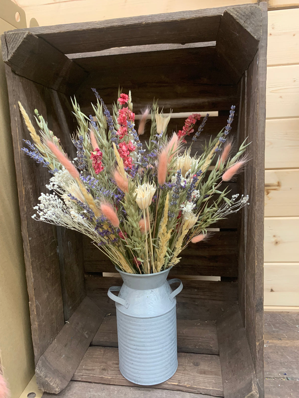 A rustic dried flower bouquet in metal milk canister, displayed inside a dark wooden crate against a light wood panel background.