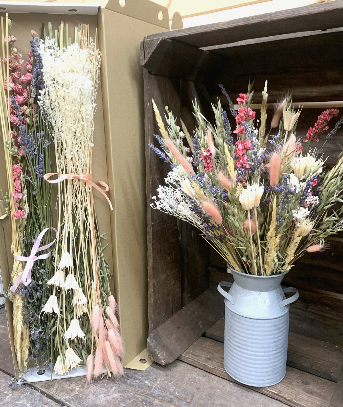 A styled arrangement of dried flowers and grasses, including pink, purple, cream, green and peach, displayed in rustic containers. On the left, a bundle of dried flowers is tied with a pink ribbon and held in a cardboard box.