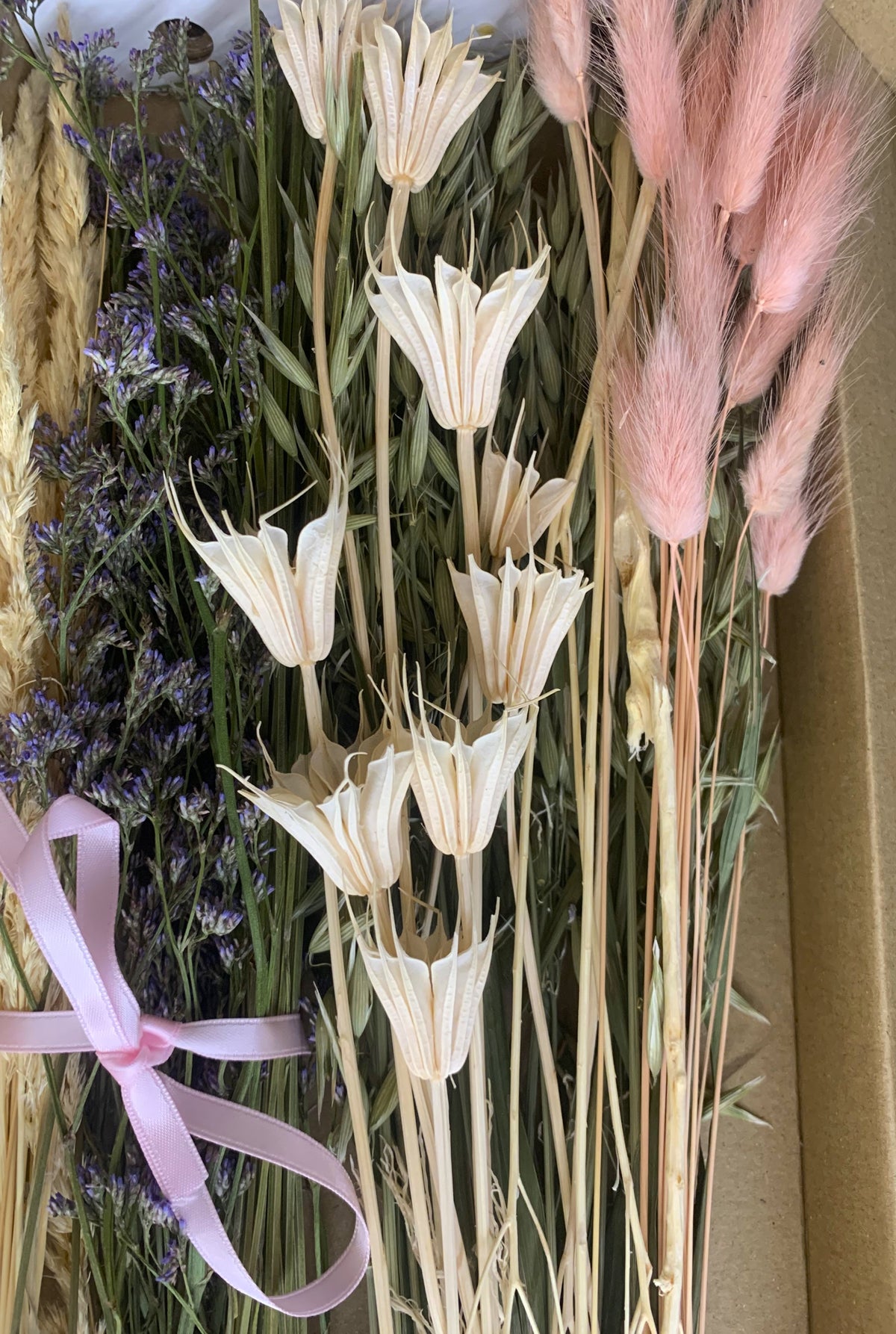 A dried flower arrangement in a brown box, featuring cream star-shaped flowers, pink bunny tails, lavender sprigs, and a light pink ribbon tied at the bottom left.