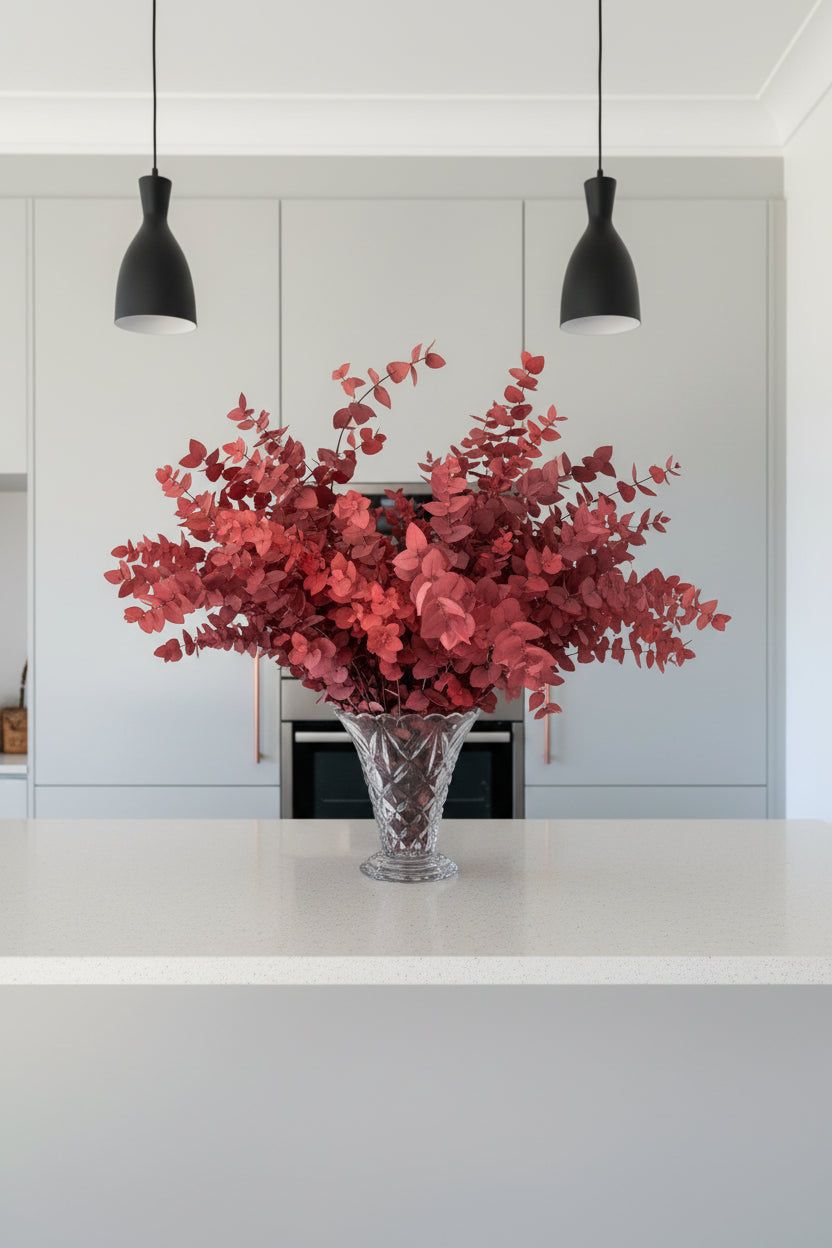 A large red leafy arrangement of preserved eucalyptus in a cut glass vase, displayed on a kitchen countertop against a grey units. 