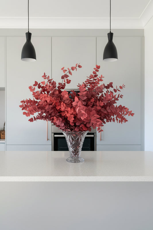 A large red leafy arrangement of preserved eucalyptus in a cut glass vase, displayed on a kitchen countertop against a grey units. 