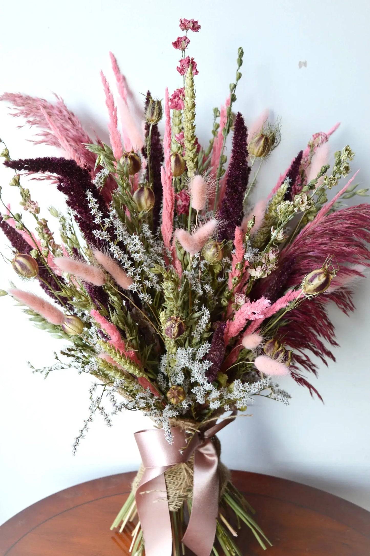 Dusky Pinks Dried Flower Gift Bouquet displayed on a wooden side table against a white wall. 