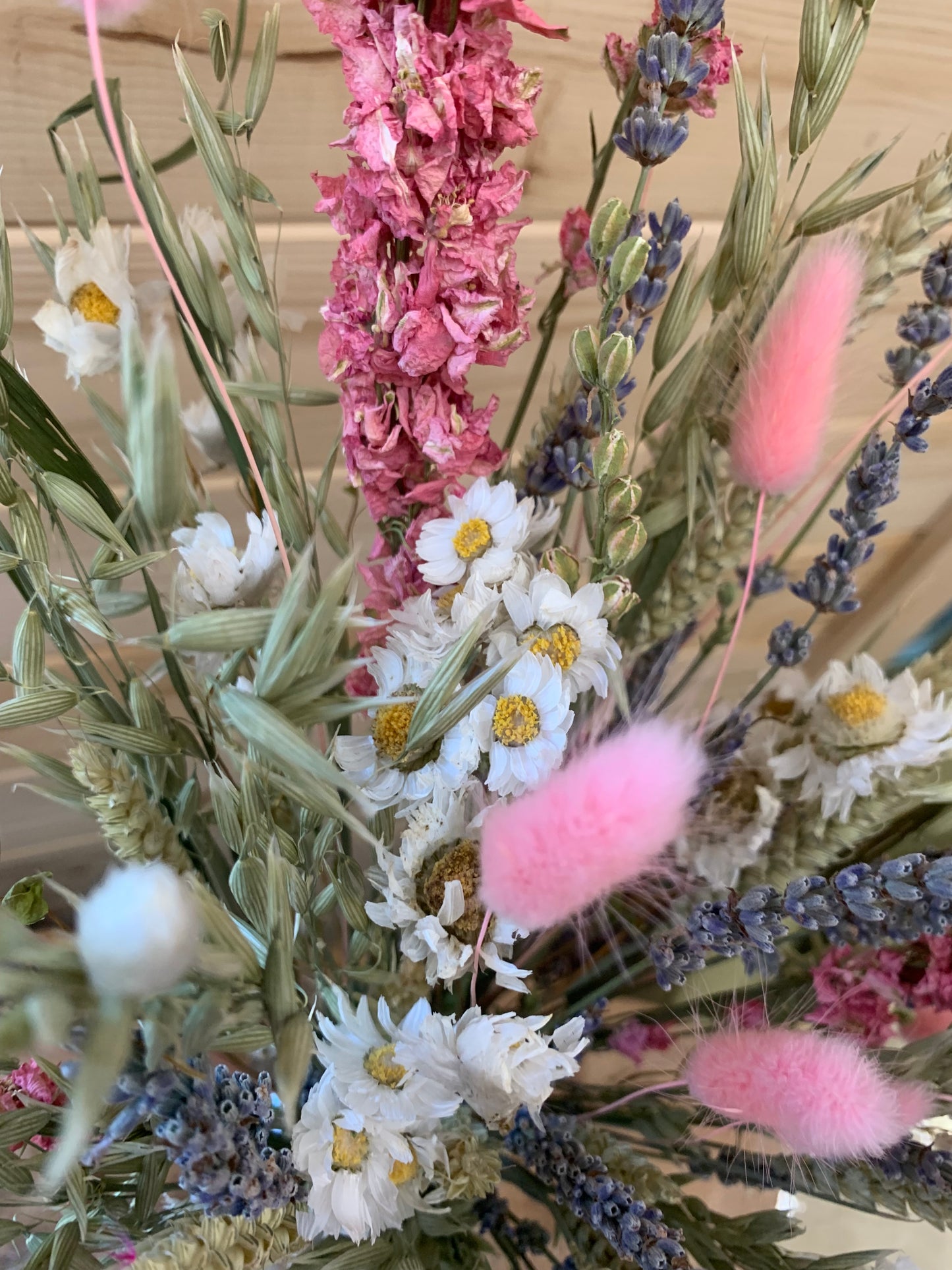 A close-up of a dried floral arrangement featuring pink fluffy bunny tails, white daisy-like flowers with yellow centers, lavender sprigs, and muted green foliage against a light wood background.