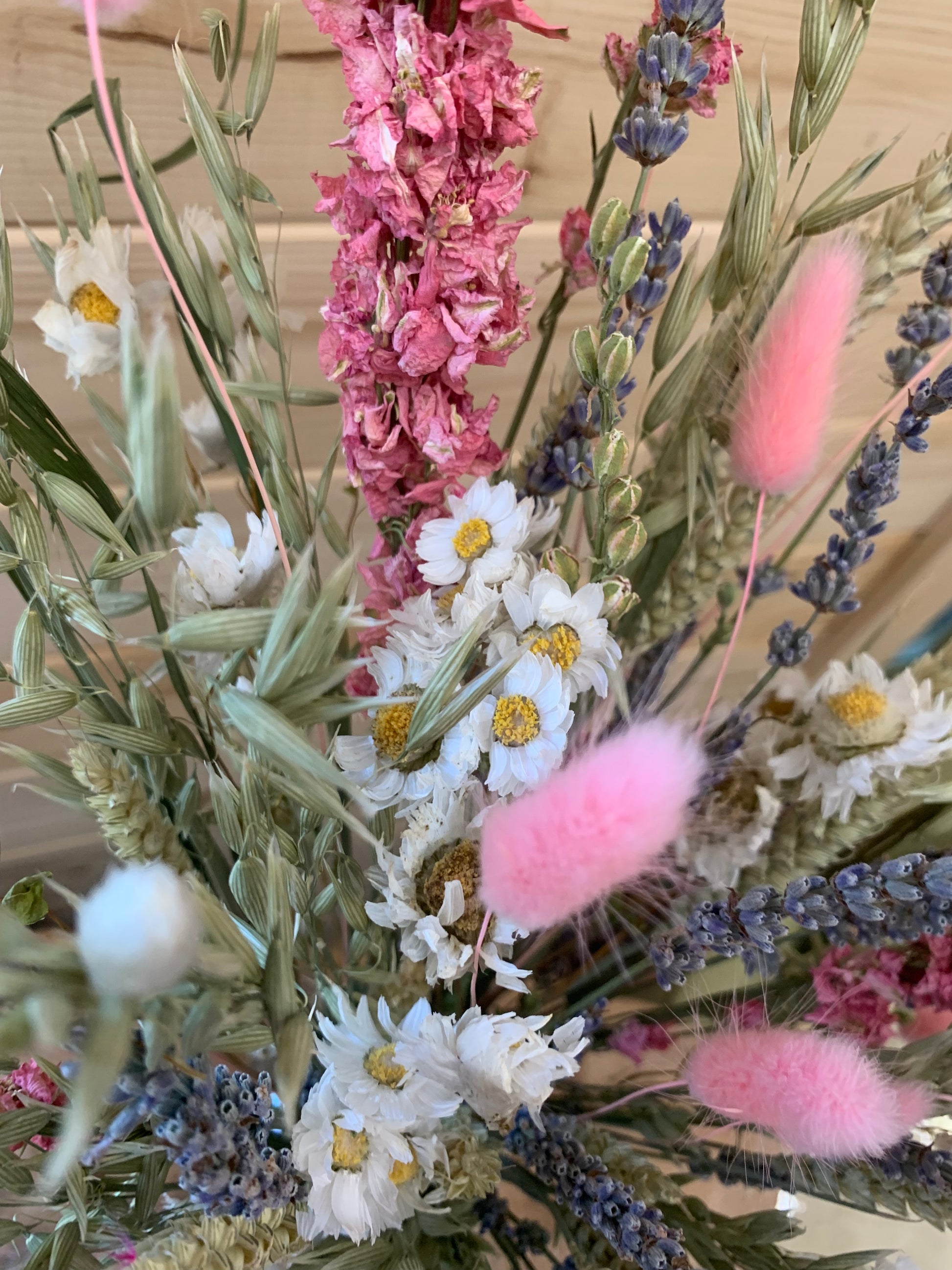 A close-up of a dried floral arrangement featuring pink fluffy bunny tails, white daisy-like flowers with yellow centers, lavender sprigs, and muted green foliage against a light wood background.