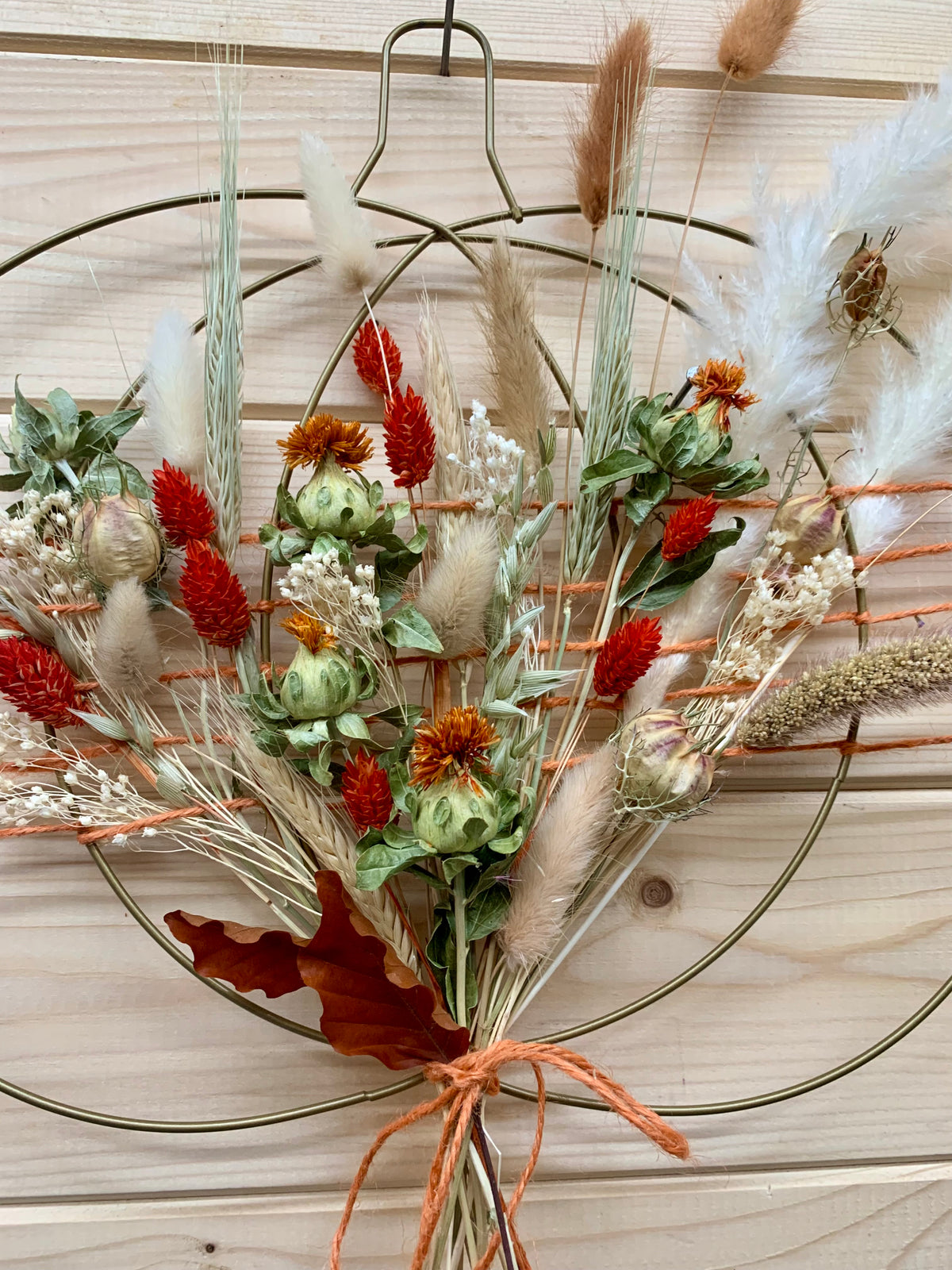 An autumn-themed dried flower wreath hanging on a light wood paneled wall. The wreath features a bronze metal circular frame filled with various dried botanicals including red amaryllis, orange poppy-like flowers, beige pampas grass, white baby's breath, and a brown leaf, all tied together with orange string.