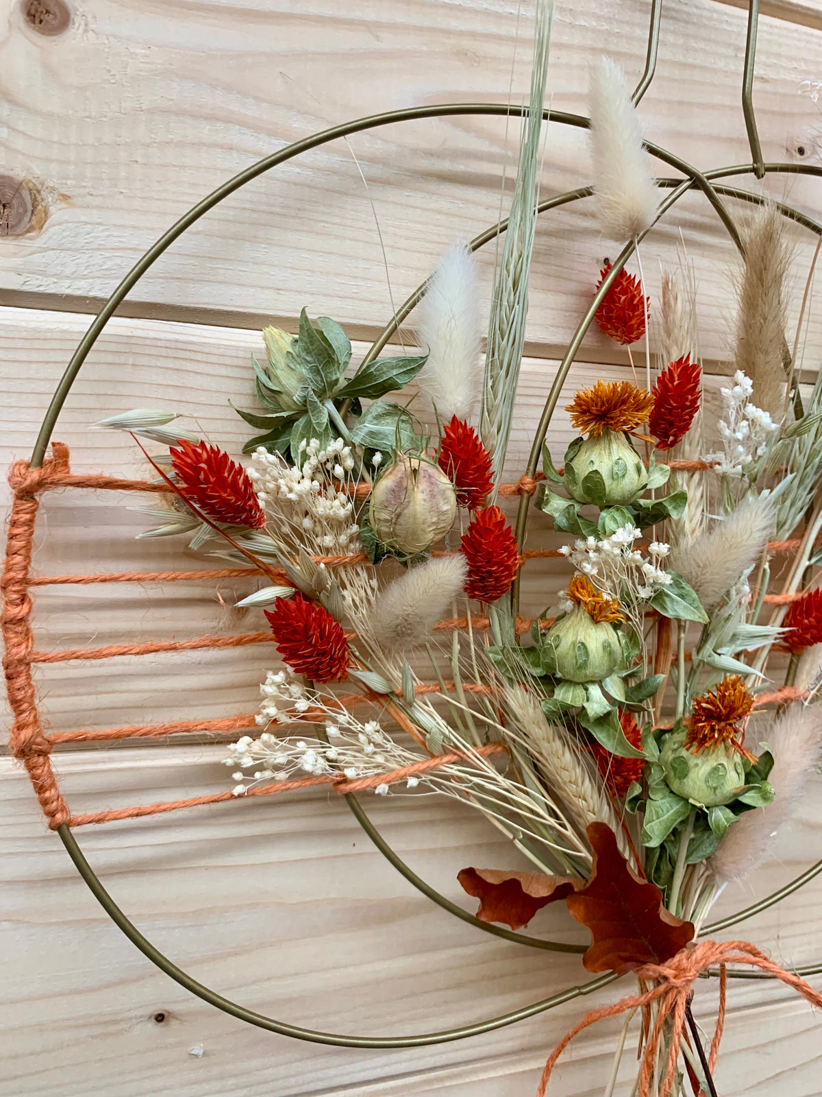 An autumn-themed wire wreath with dried grasses, red flowers, and green leaves, tied with orange string against a light wood plank background.