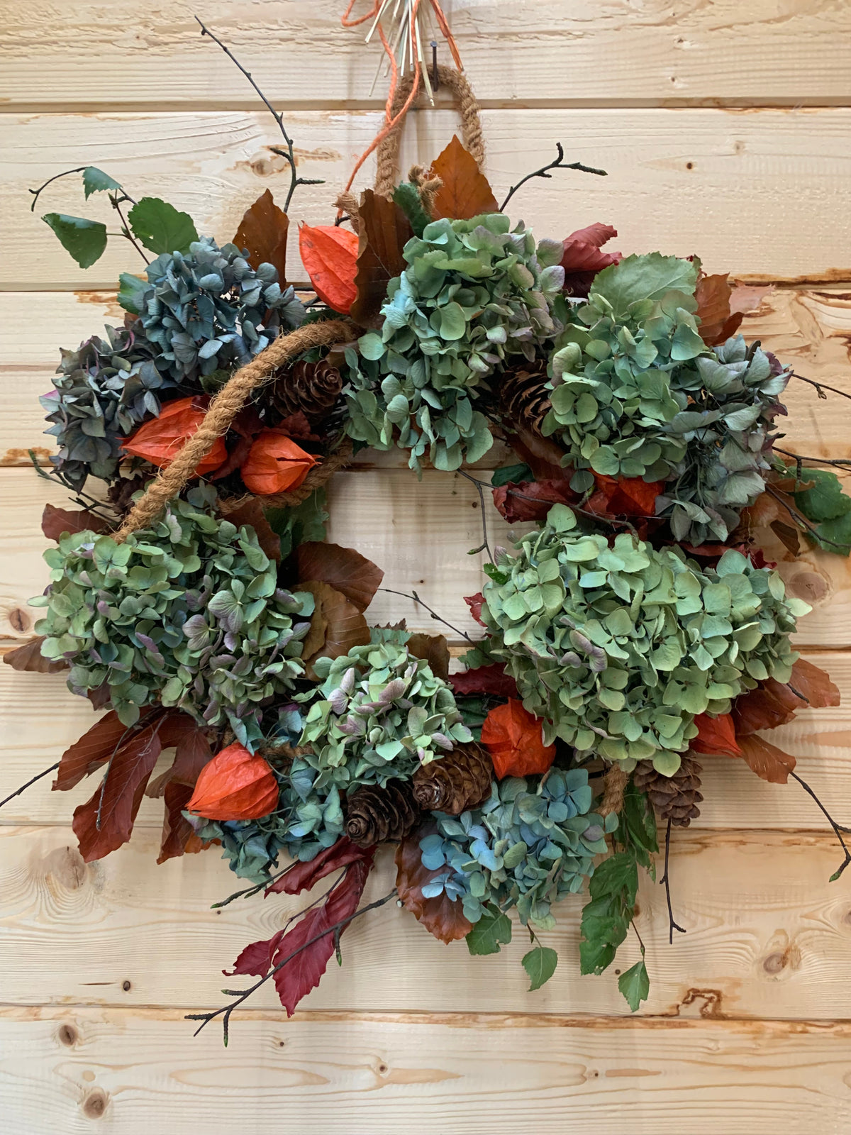 A rustic autumn wreath made of dried green hydrangea blooms, orange Chinese lanterns, pinecones, brown leaves, and a natural jute rope handle, hanging against a light wood plank wall.