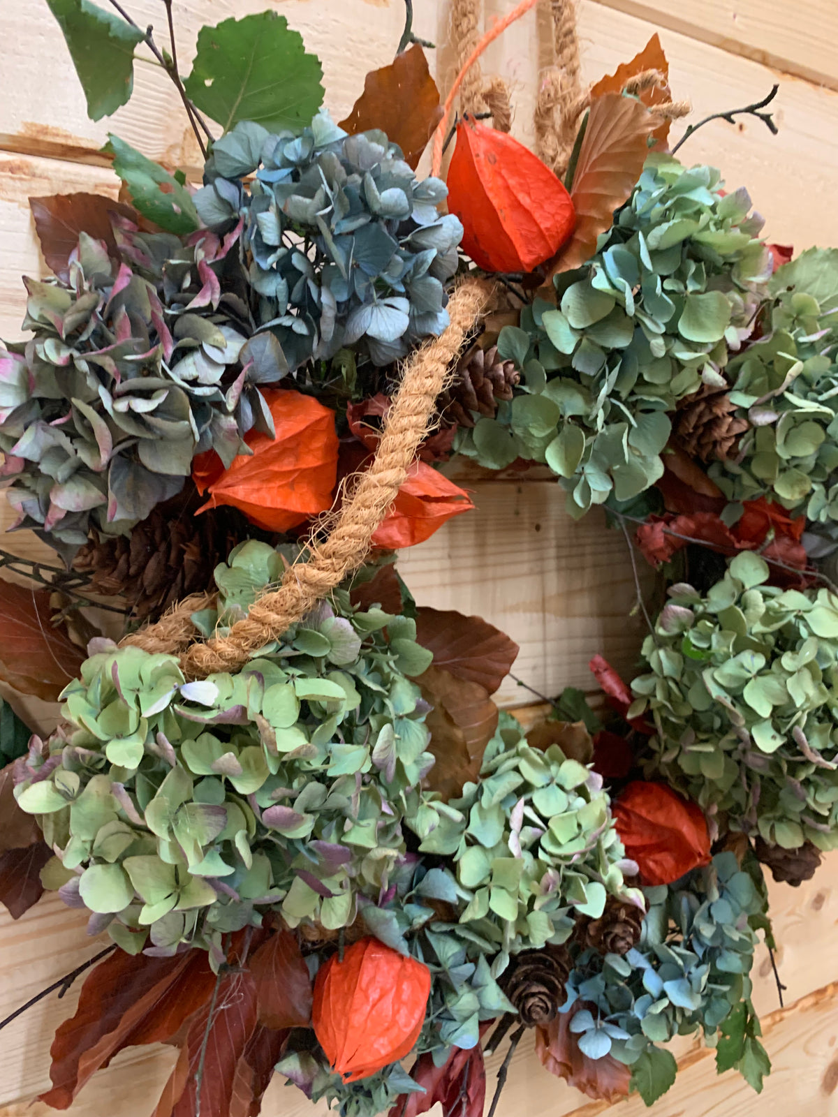 An autumn-themed dried flower wreath featuring muted green and blue-gray hydrangeas, orange lanterns, pinecones, and brown leaves, hanging on a light wood paneled wall.