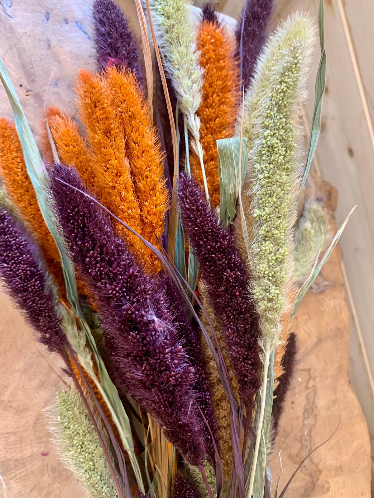 A close-up of a dried floral arrangement featuring tall ornamental grasses in vibrant orange, deep burgundy-purple, and muted sage green, set against a rustic wood background.