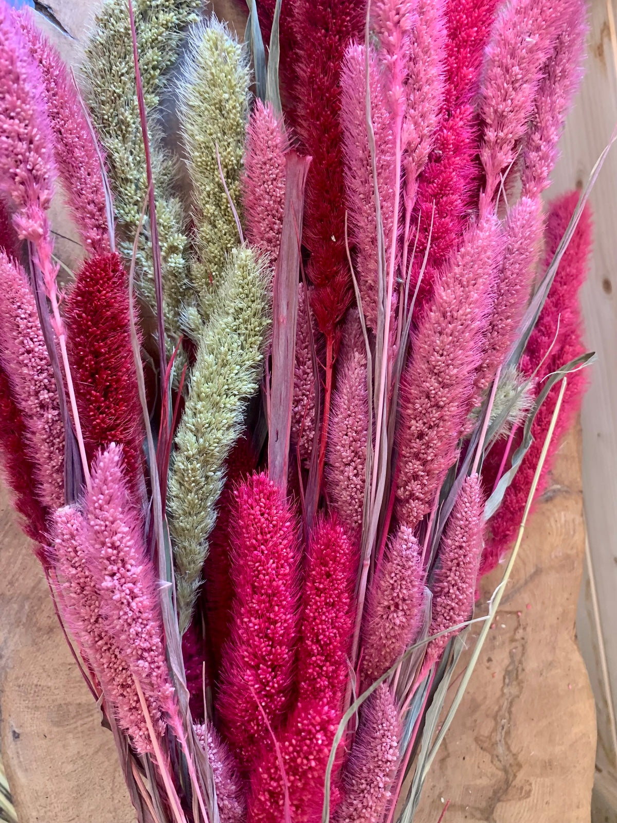 A close-up of dried ornamental grasses in vivid pink, magenta, and sage green tones, arranged against a rustic wood background.