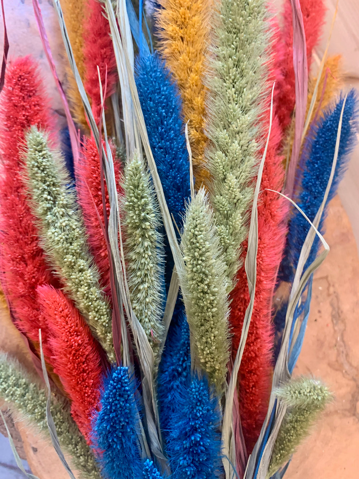 A close-up of a dried floral arrangement featuring fluffy decorative grasses in bright red, blue, sage green, and mustard yellow tones, with pale straw stems and leaves.