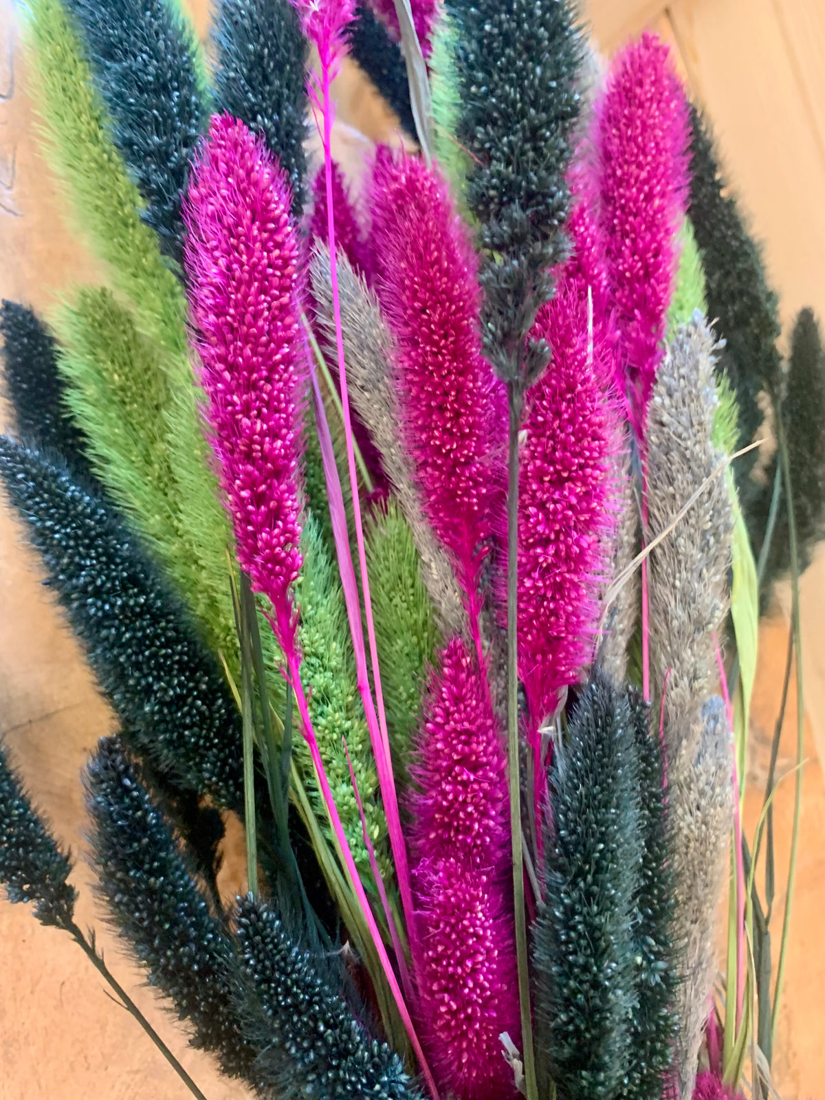 A close-up of a dried floral arrangement featuring vibrant magenta, lime green, and dark charcoal fluffy grass spikes on thin green stems.